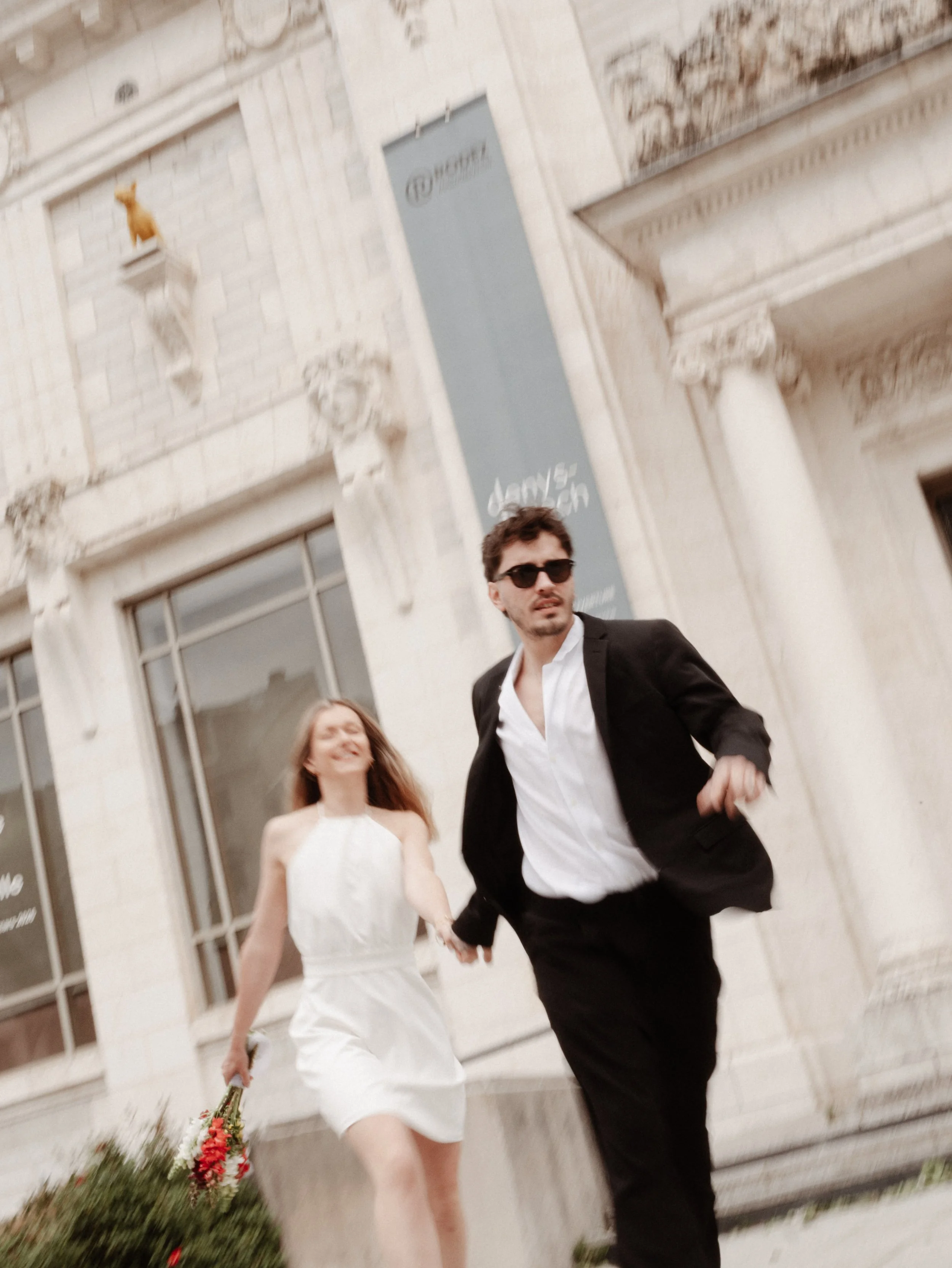 Jeune couple se tenant la main en marchant devant un bâtiment en pierre avec une statue de chat sur une plateforme. L'homme porte un costume noir avec chemise blanche et lunettes de soleil, la femme porte une robe blanche et tient un bouquet de fleur