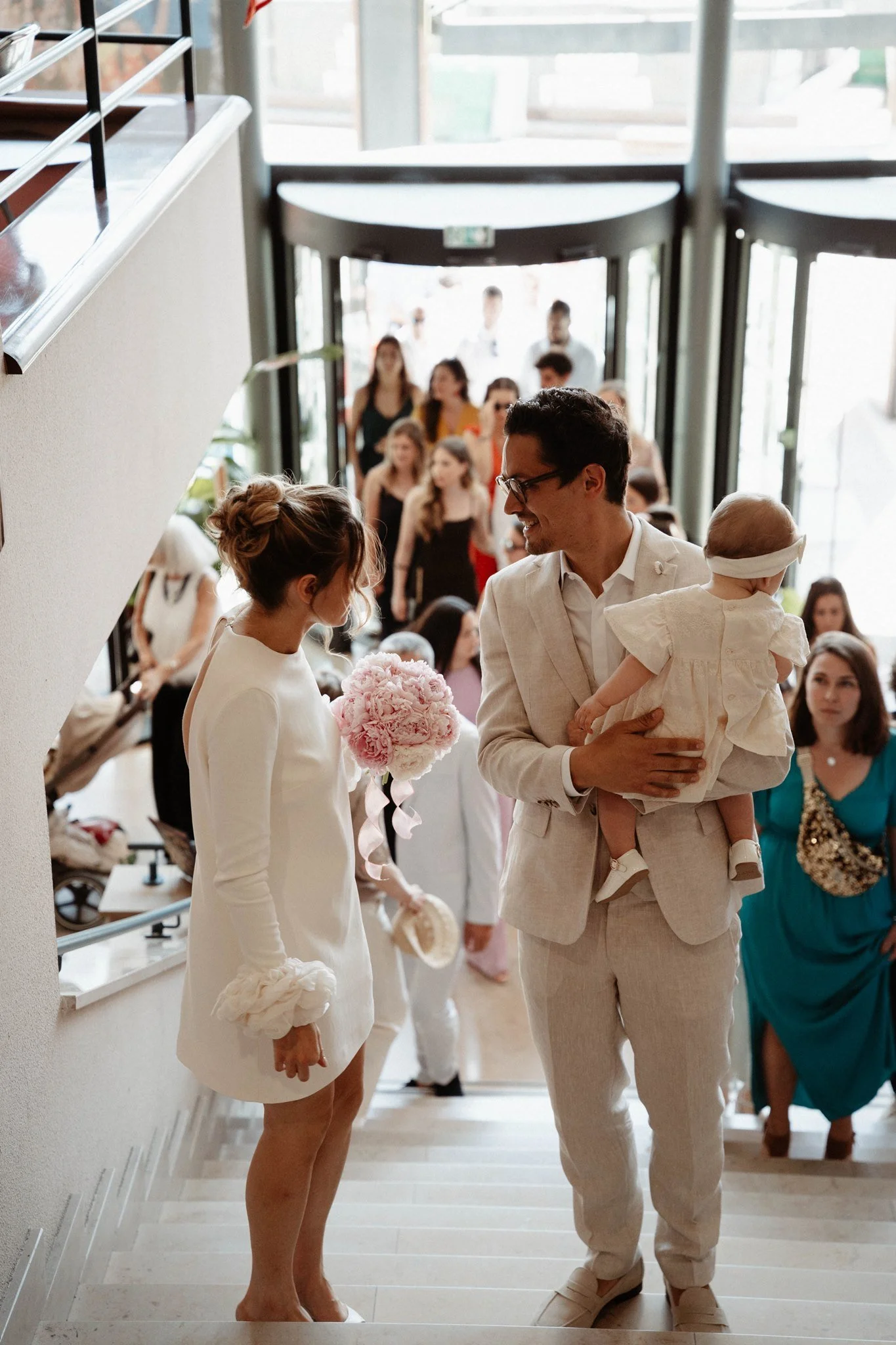 Un homme et une femme habillés en blanc dans une ambiance de mariage, la femme tient un bouquet de fleurs roses, ils semblent échanger un regard. Autour, plusieurs personnes en tenue élégante participent à la célébration.