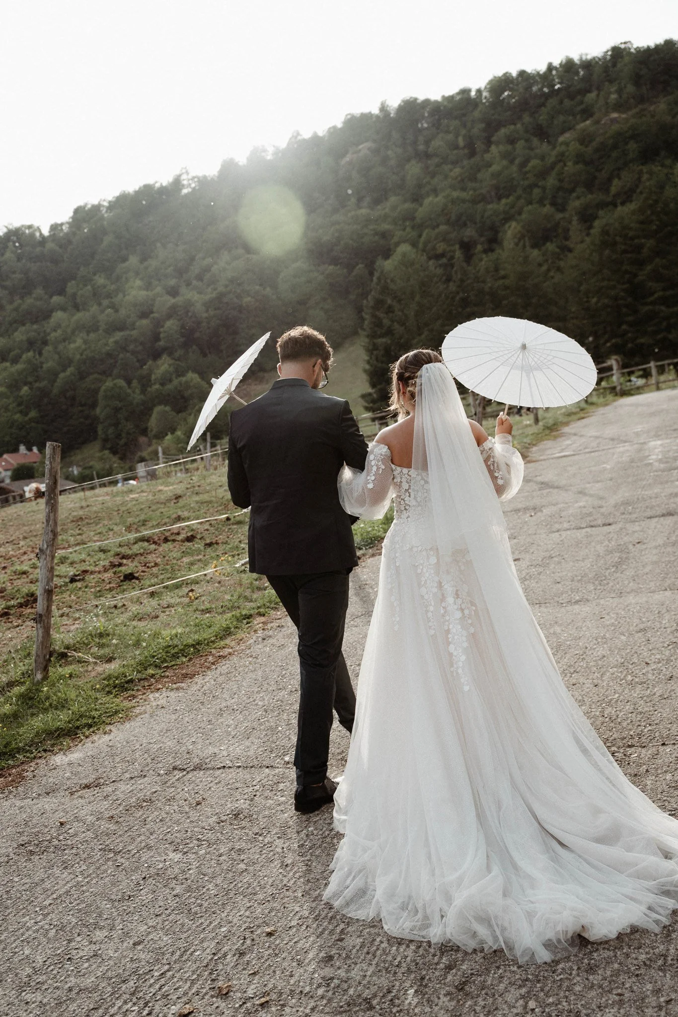 Un couple en mariage marchant avec des parapluies blancs dans une campagne avec des collines verdoyantes en arrière-plan.
