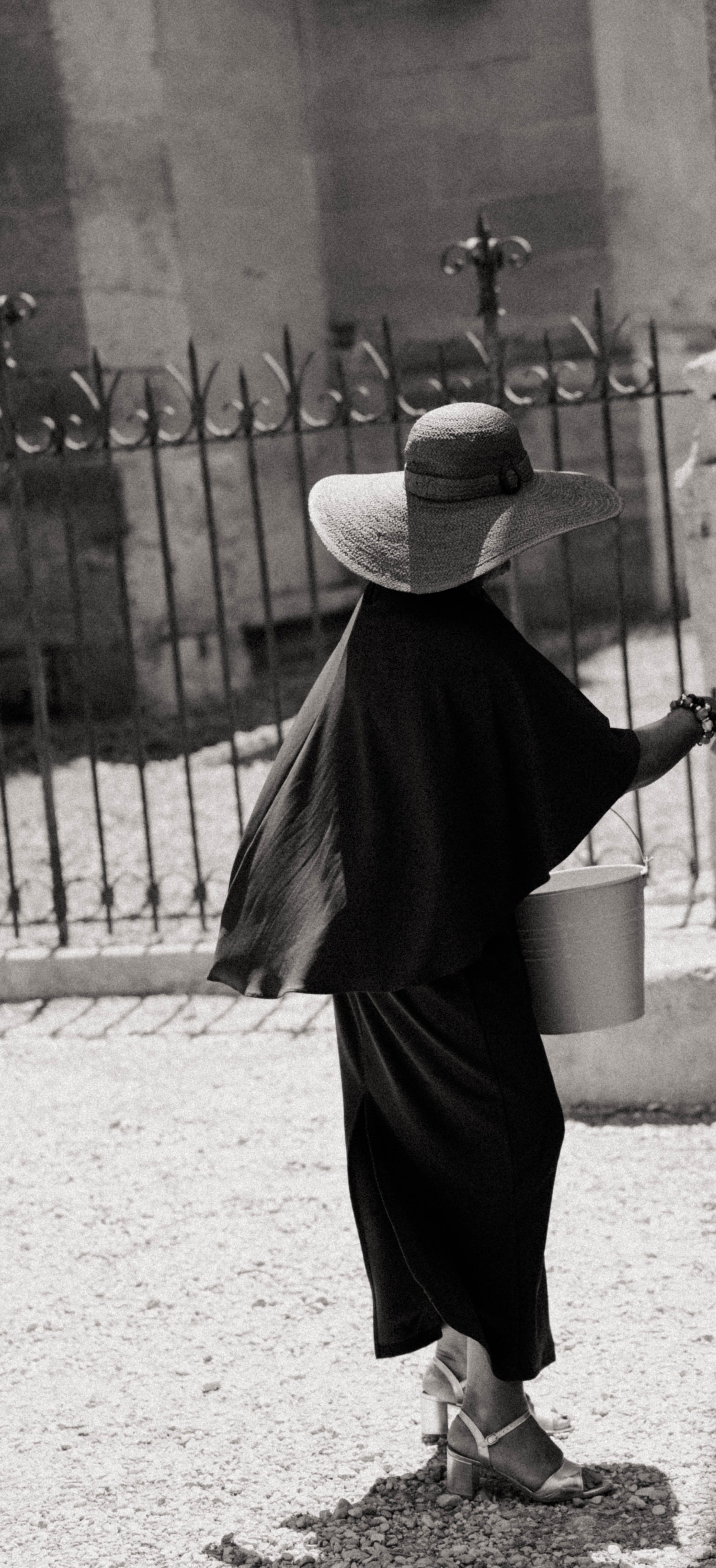 Femme portant un grand chapeau de paille et une robe noire, tenant un seau, marchant devant une grille en fer forgé fanée, en noir et blanc.