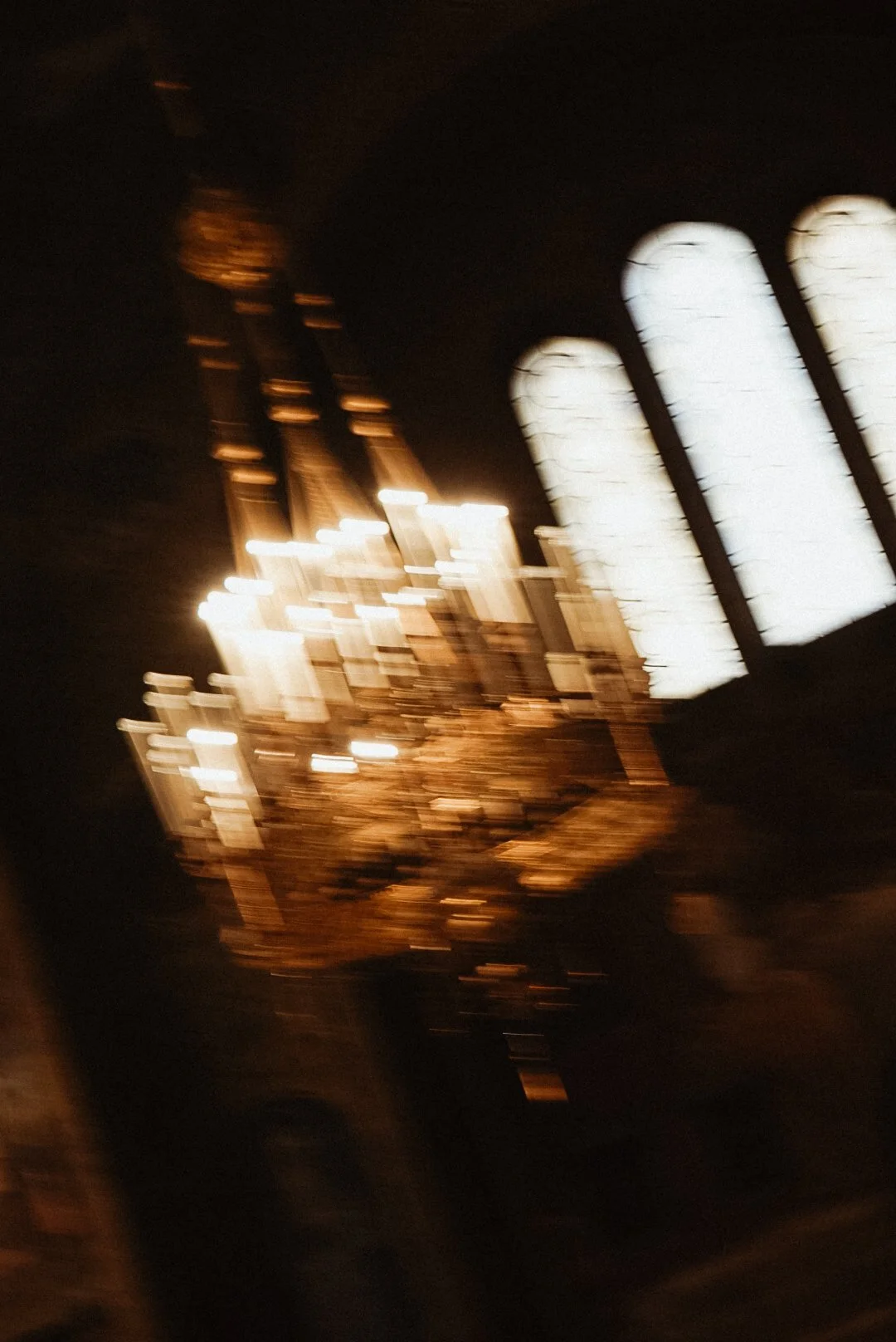 Image floue montrant un lustre en cristal avec des lampes allumées et une fenêtre en forme d'arche dans un environnement sombre.