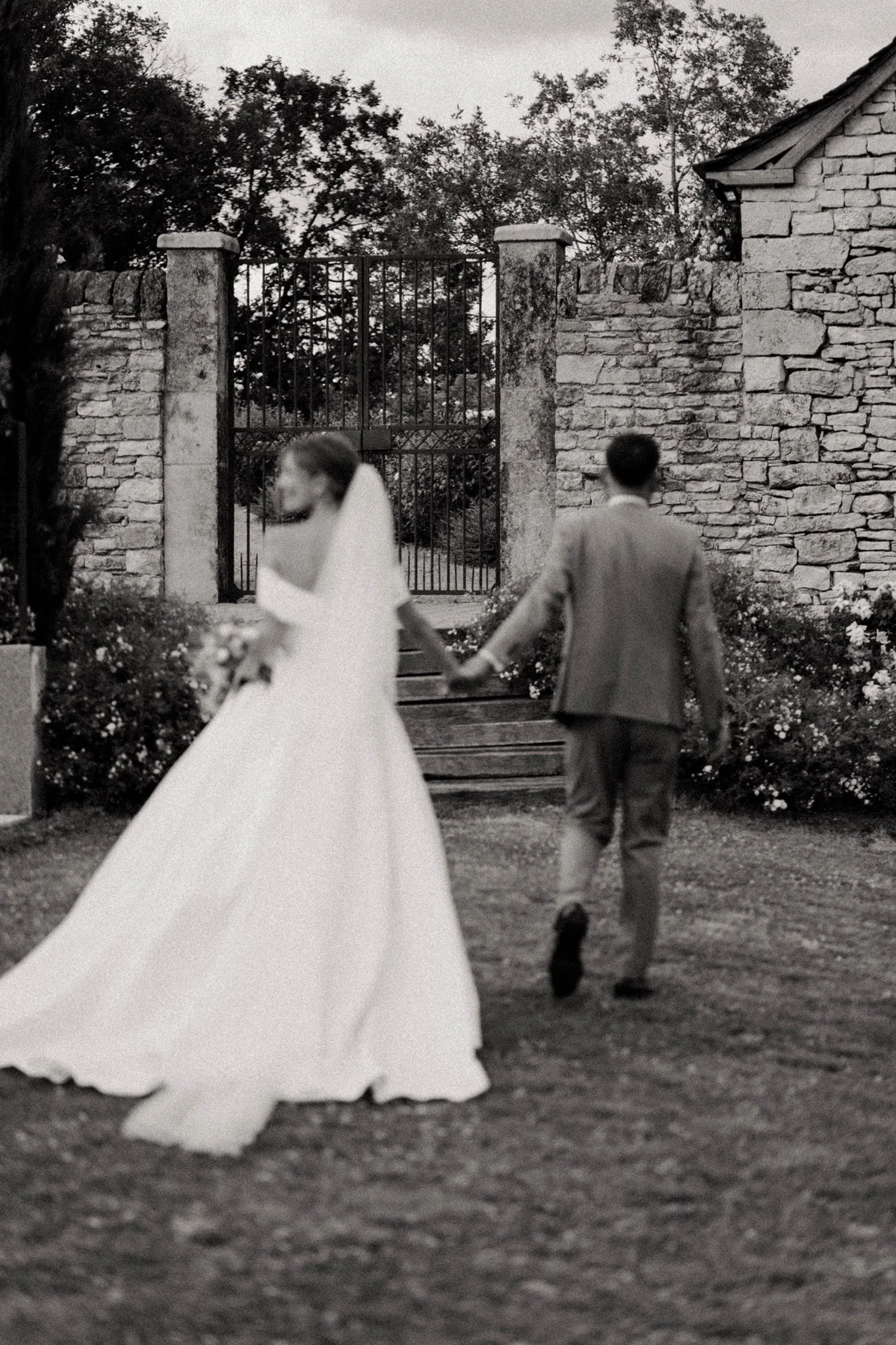 Un couple marchant main dans la main devant une clôture en pierre et un portail en fer forgé, dans un jardin avec des arbres et des fleurs, en noir et blanc.