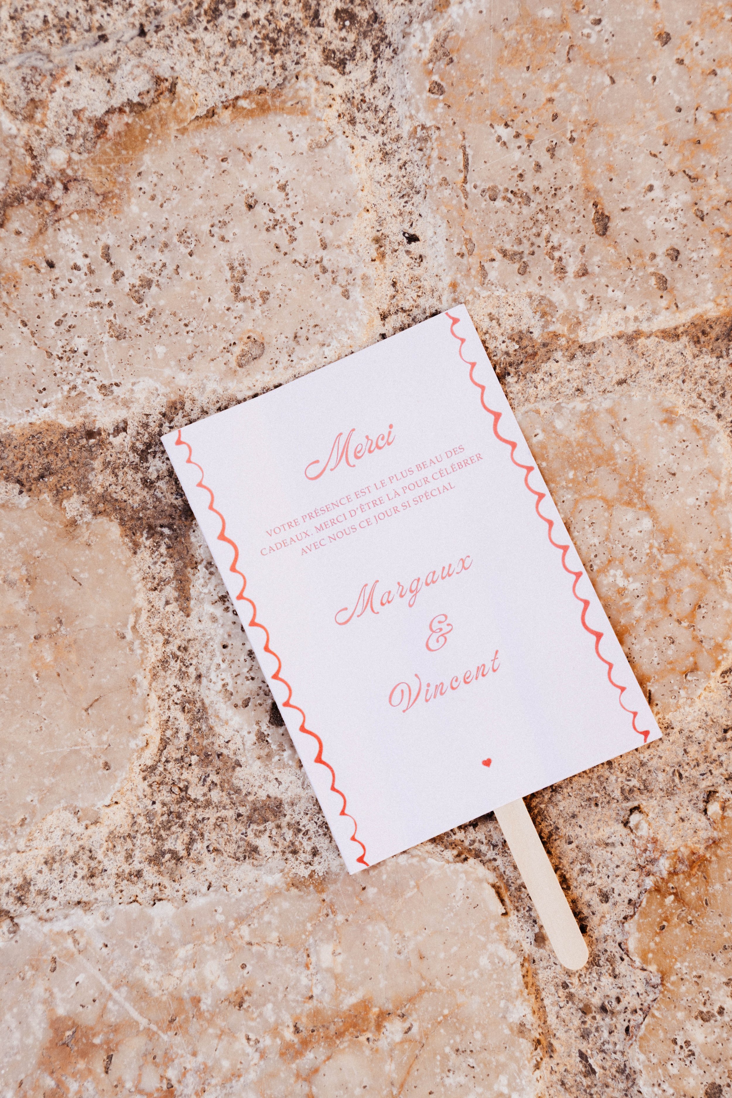 Carte de remerciement en papier blanc avec une bordure ondulée orange, posée sur une surface en pierre beige. La carte contient un message en français pour un mariage.