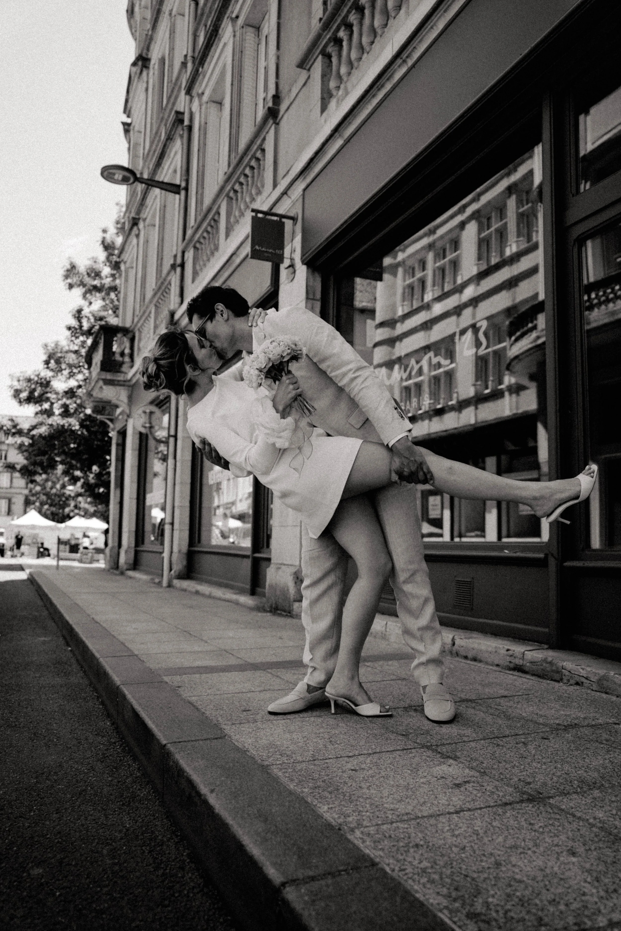 Un couple en mariage s'embrasse et partage un baiser passionné dans la rue, l'homme en costume beige et la femme en robe blanche, entourés d'une architecture urbaine et de bâtiments en arrière-plan.