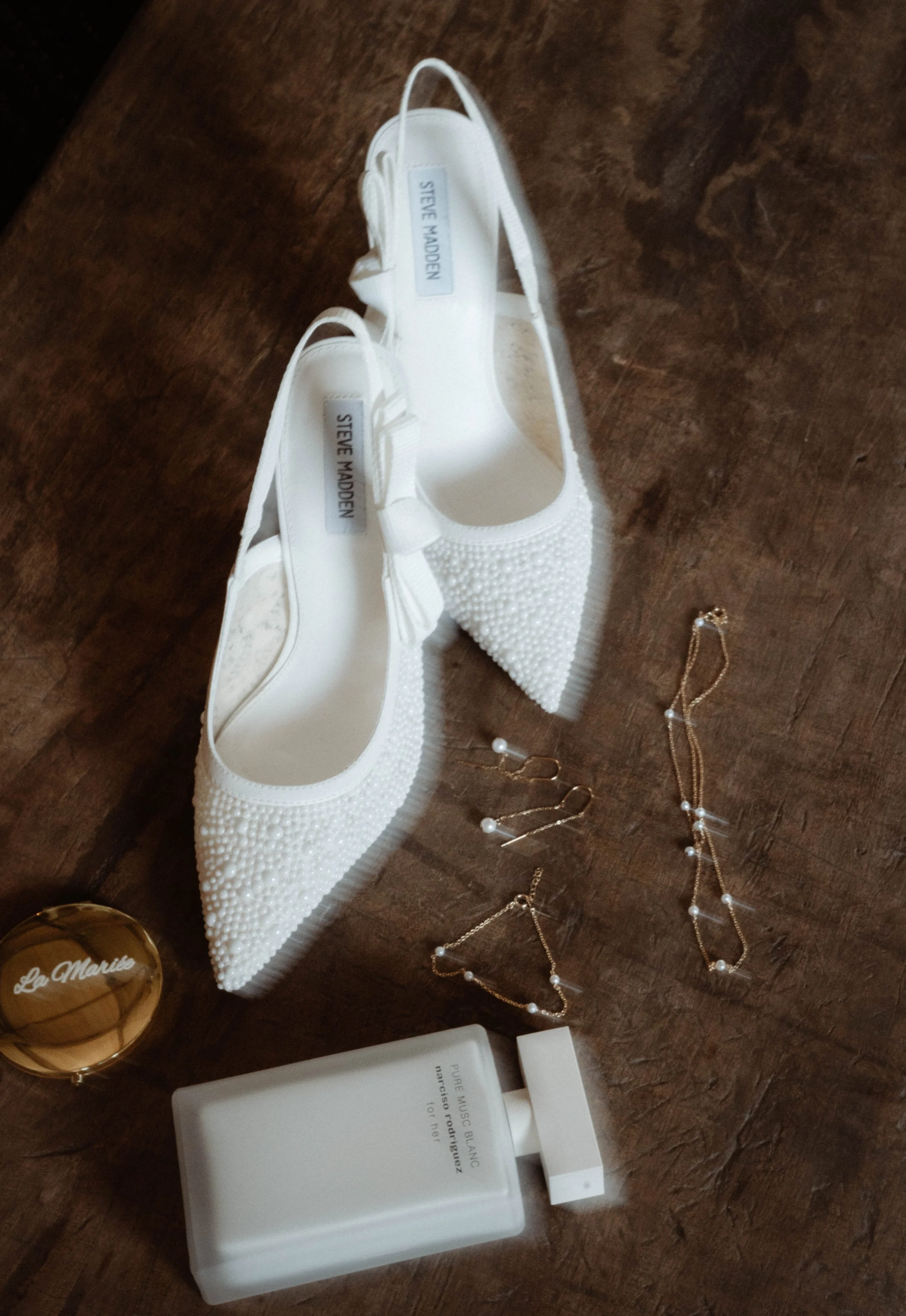 Chaussures blanches décorées de perles, accessoires de bijouterie en or avec perles, un parfum blanc, et un dictionnaire blanc sur une surface en bois.