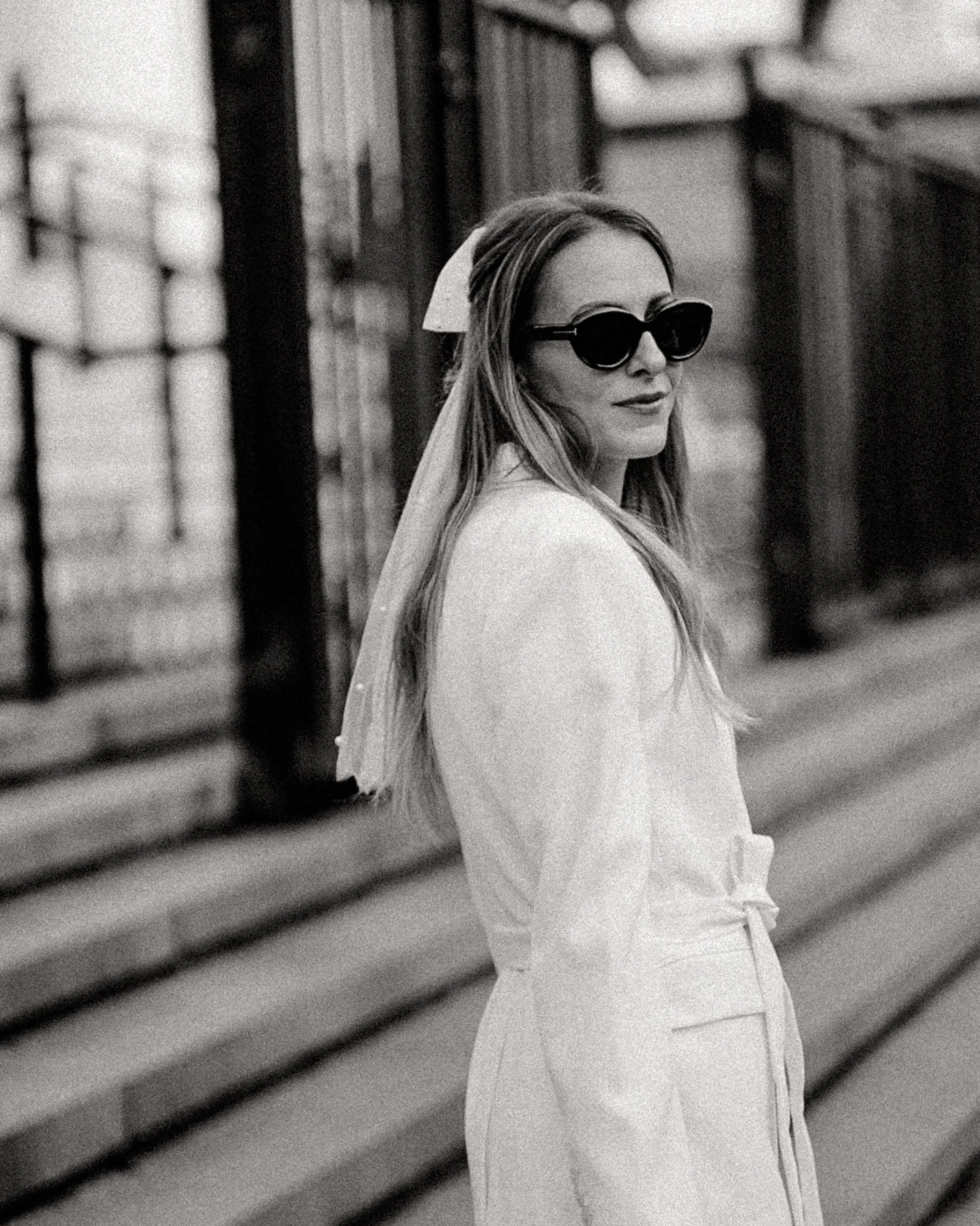 Une femme porte des lunettes de soleil foncées, une tenue blanche et un foulard blanc, debout près d'une grille en métal dans une rue