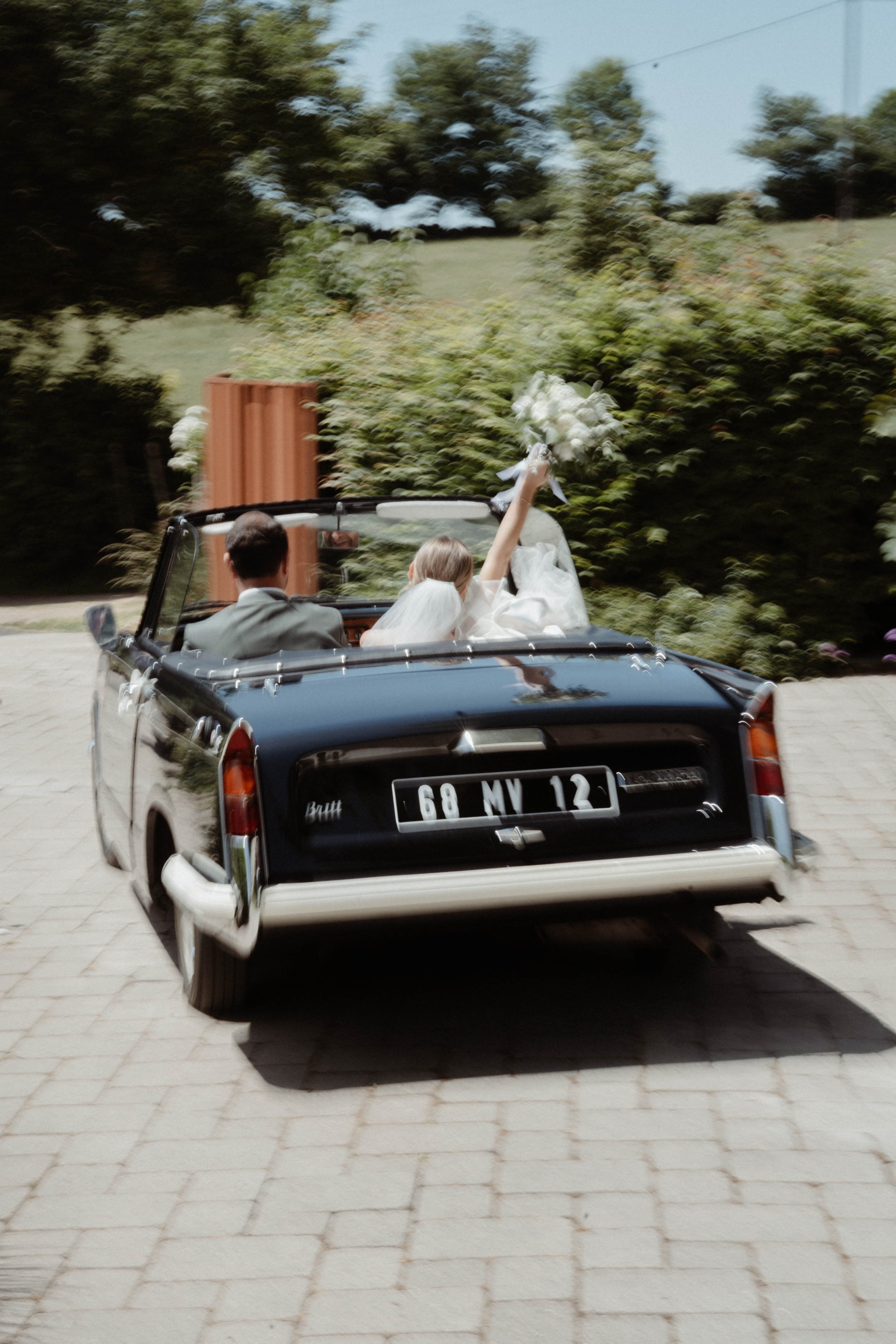 Voiture de mariage noire avec un couple à l'intérieur, avec la femme levant ses fleurs, en mouvement sur un chemin pavé, entouré de végétation.