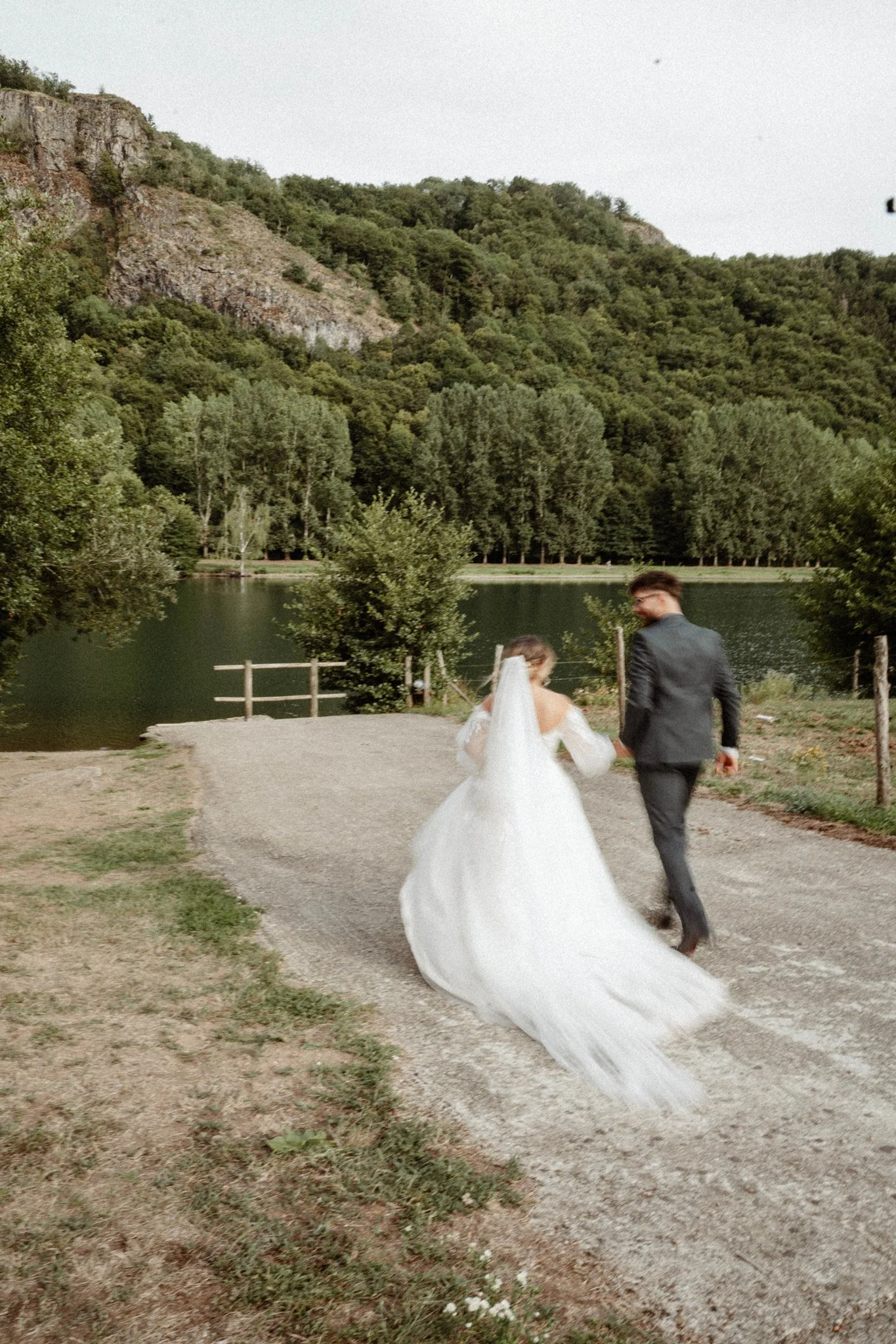 Un couple vêtu de vêtements de mariage marche près d'un lac entouré de montagnes et d'arbres lush. La femme porte une robe de mariée blanche avec un voile long, et l'homme porte un costume sombre. La scène est en extérieur, pendant la journée, avec u