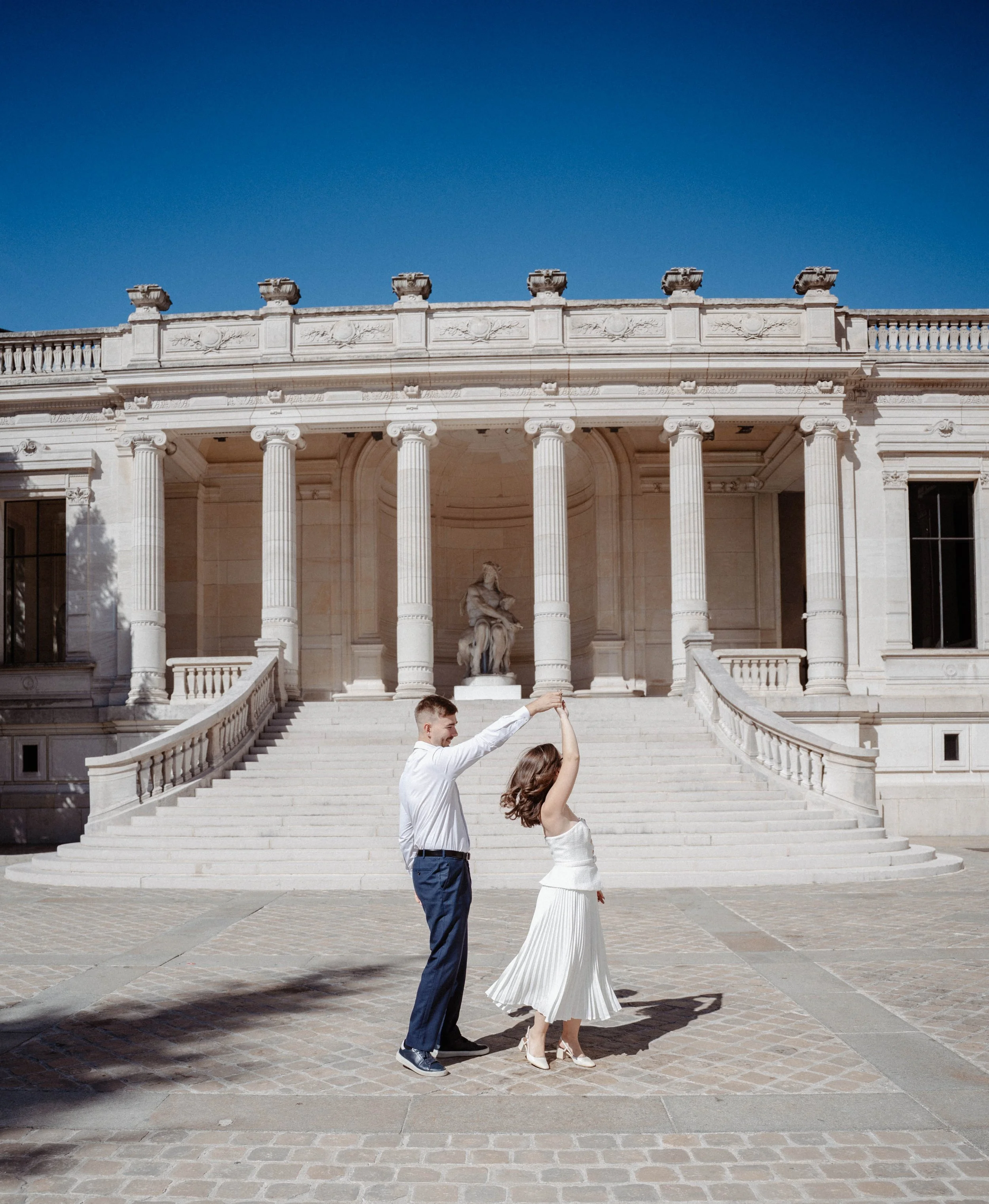 Un couple danse devant un bâtiment blanc, avec une femme en robe blanche et un homme en pantalon bleu et chemise blanche. Ils se tiennent par la main dans un style de danse en couple, devant de grands escaliers et une statue de femme au centre.