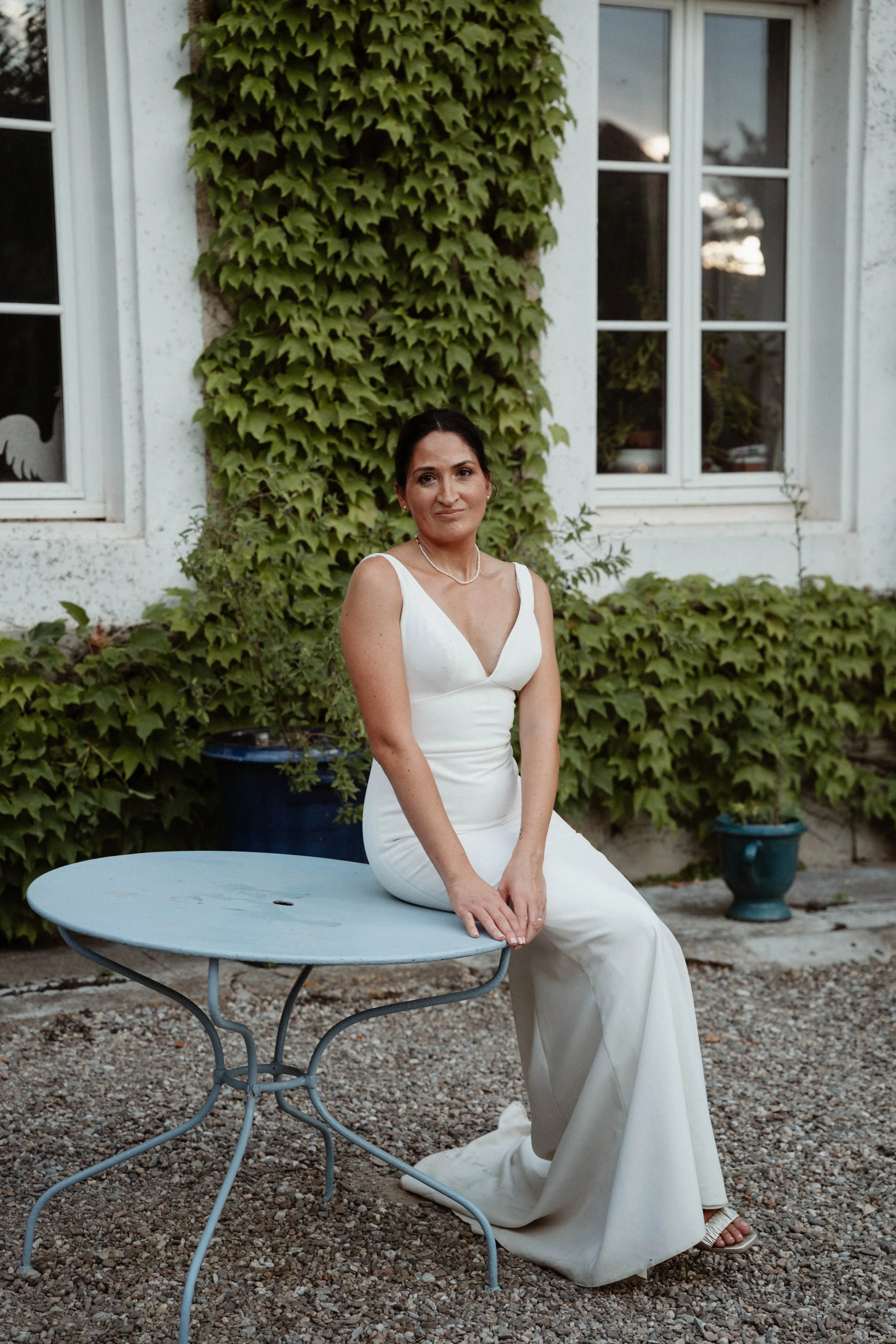 Une femme portant une robe blanche assise sur une table en métal dans un jardin. Arbres et une maison en arrière-plan.