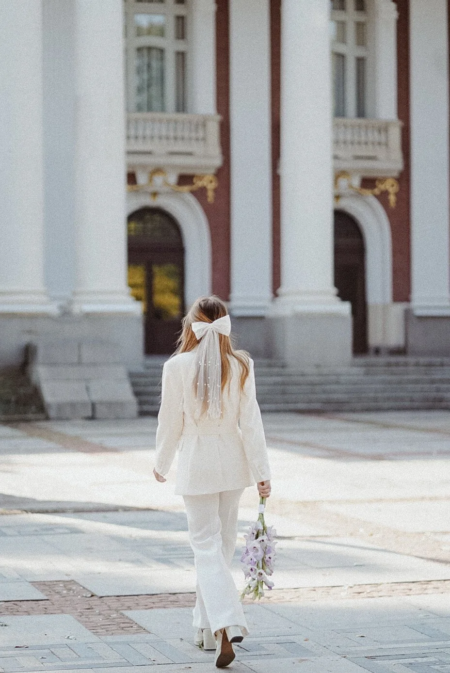 Une jeune femme habillée en blanc avec un grand nœud blanc dans les cheveux, tenant un bouquet de fleurs, marchant vers un bâtiment avec de grandes colonnes.