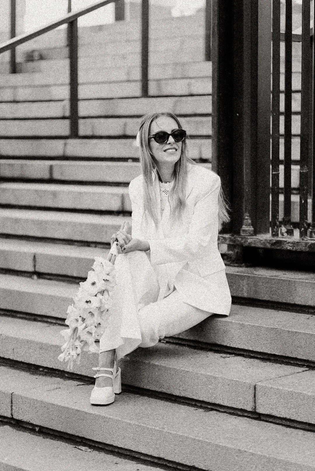 Une femme assise sur des escaliers en pierre, portant des lunettes de soleil sombres, un costume blanc, et tenant un bouquet de fleurs. Elle sourit en regardant légèrement à droite.