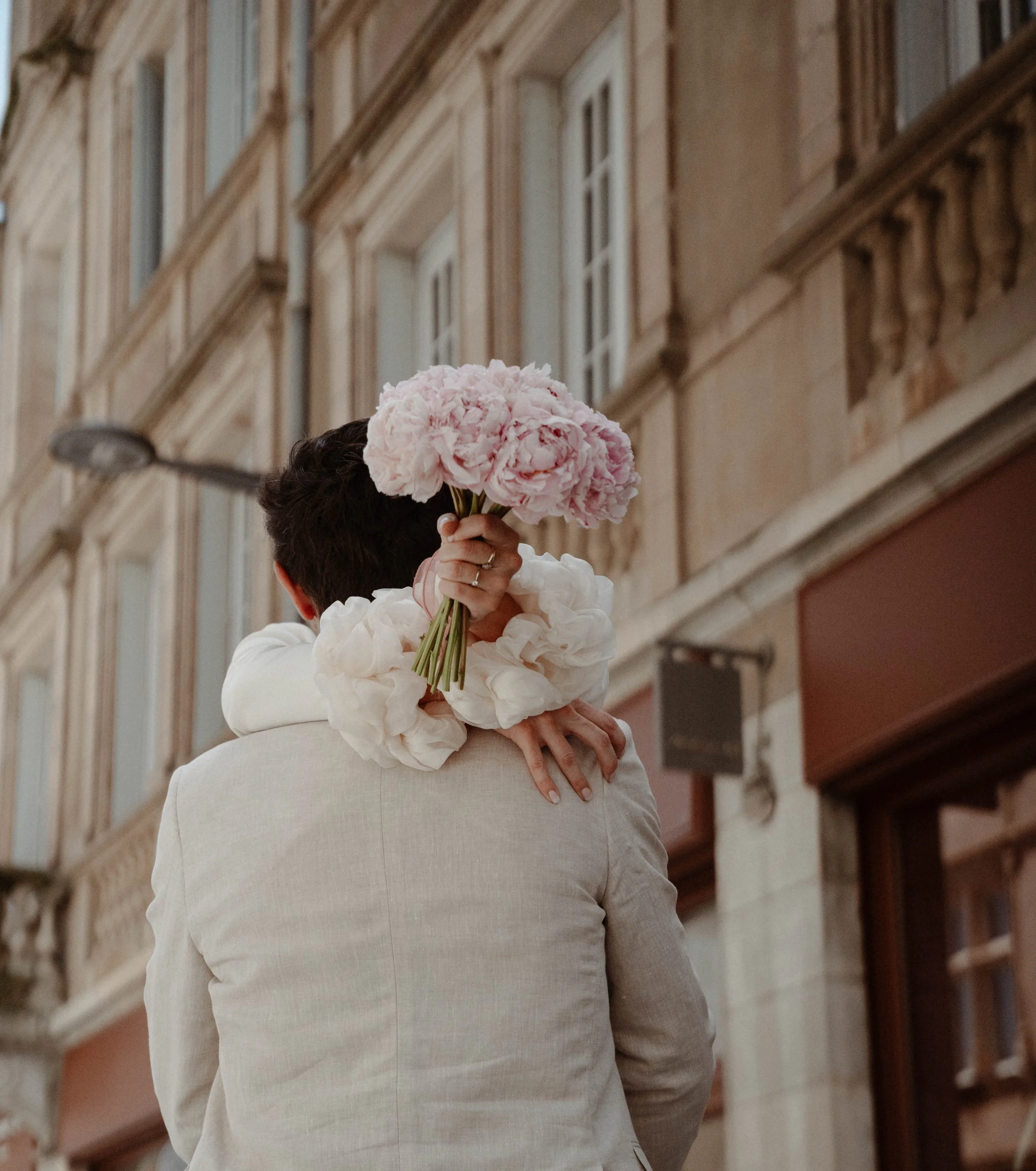Personne tenant un bouquet de fleurs roses dans une rue pavée, avec un bâtiment ancien en arrière-plan.