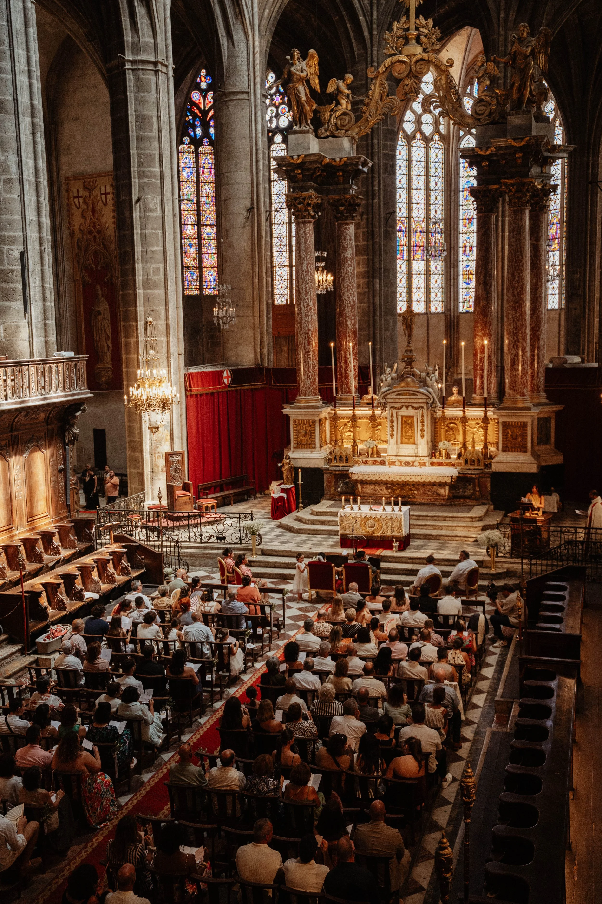 L'intérieur d'une église gothique avec un groupe de personnes assises, un autel au centre, de hautes vitres colorées et des colonnes en marbre rouge.