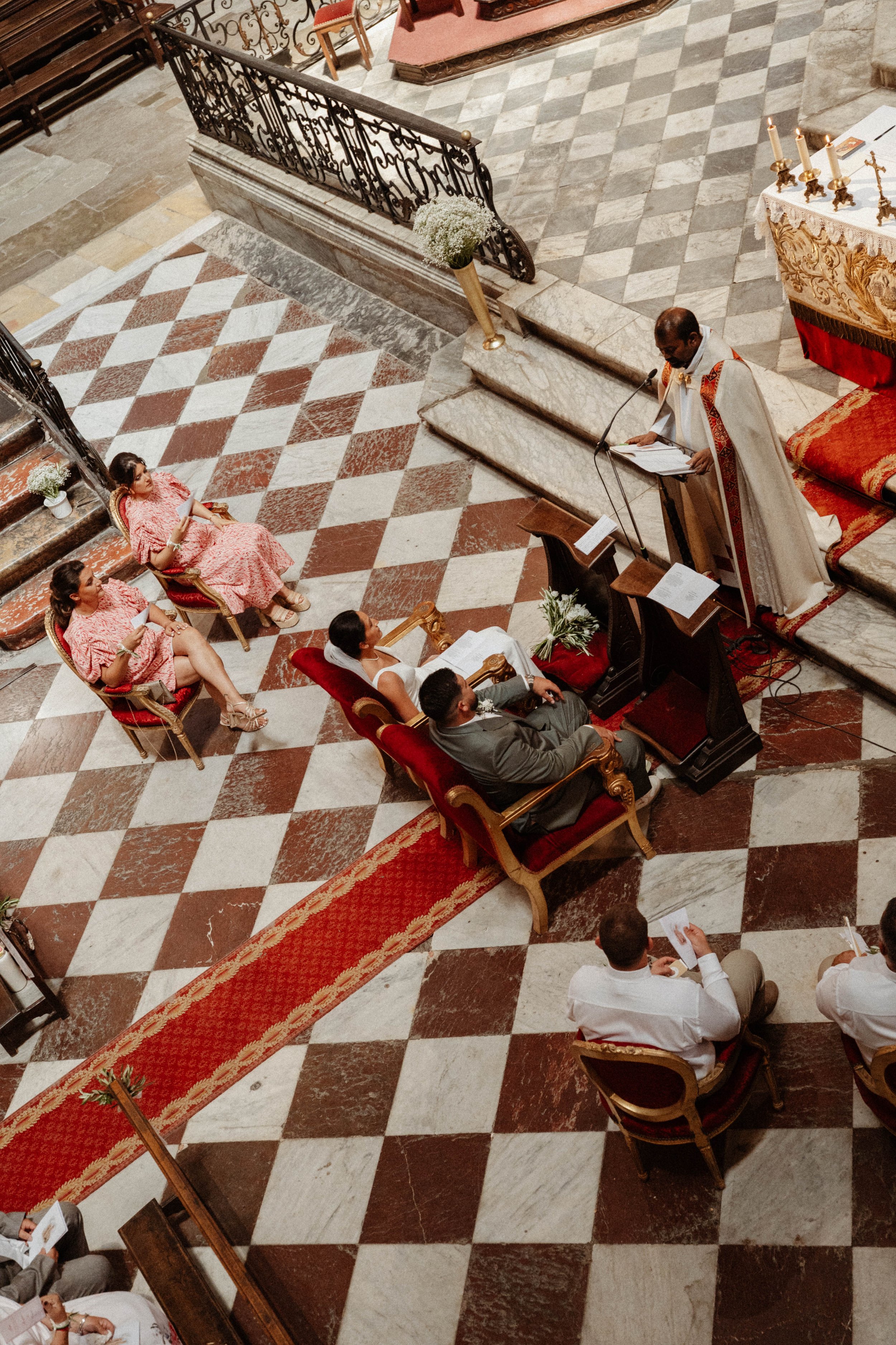 Vue en plongée d'une cérémonie religieuse dans une église, des personnes assises en rangée, un prêtre à l'autel, chandeliers et décorations religieuses.