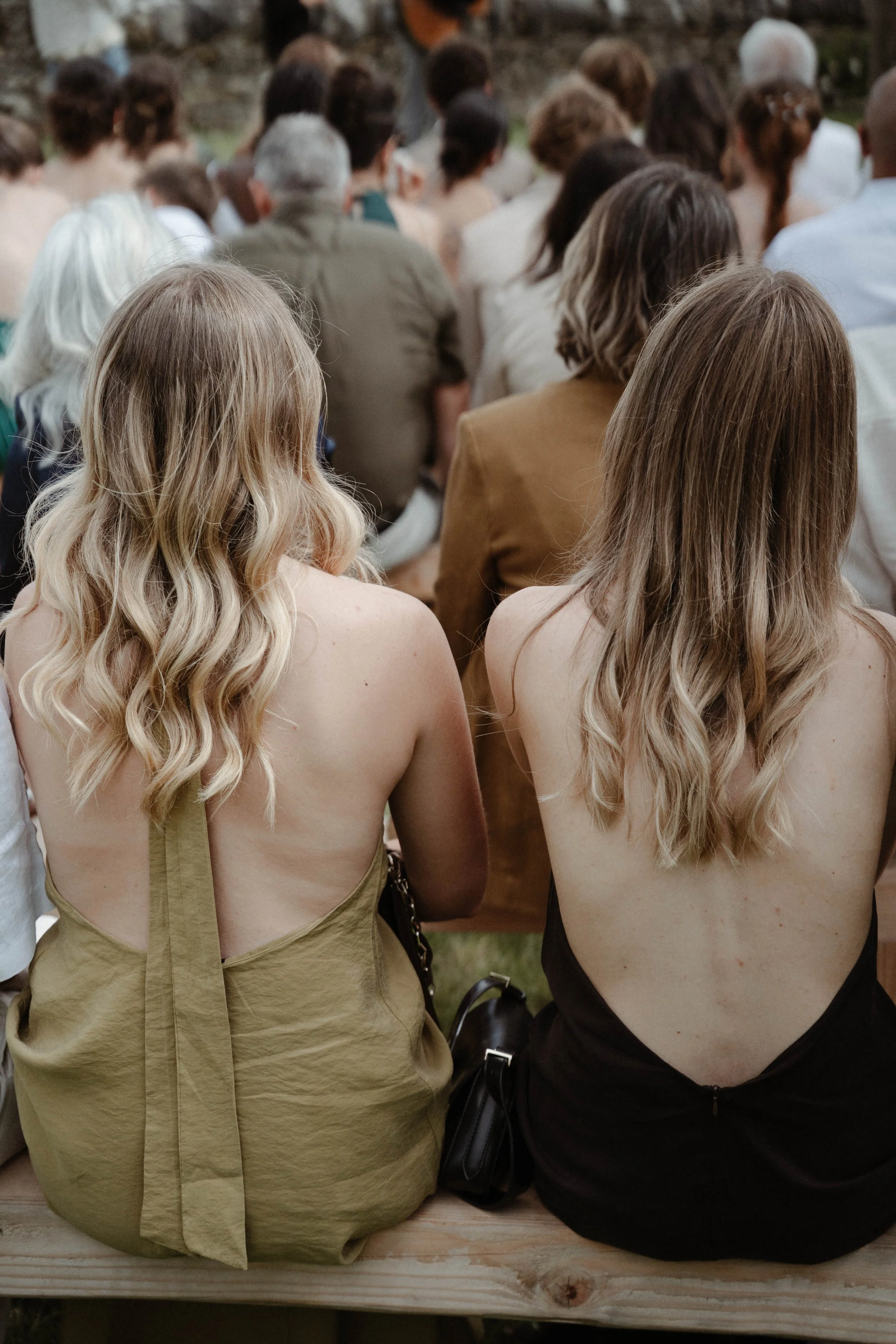 Deux femmes aux cheveux longs et ondulés, assises sur un banc en bois, vues de dos, lors d'un événement en plein air.