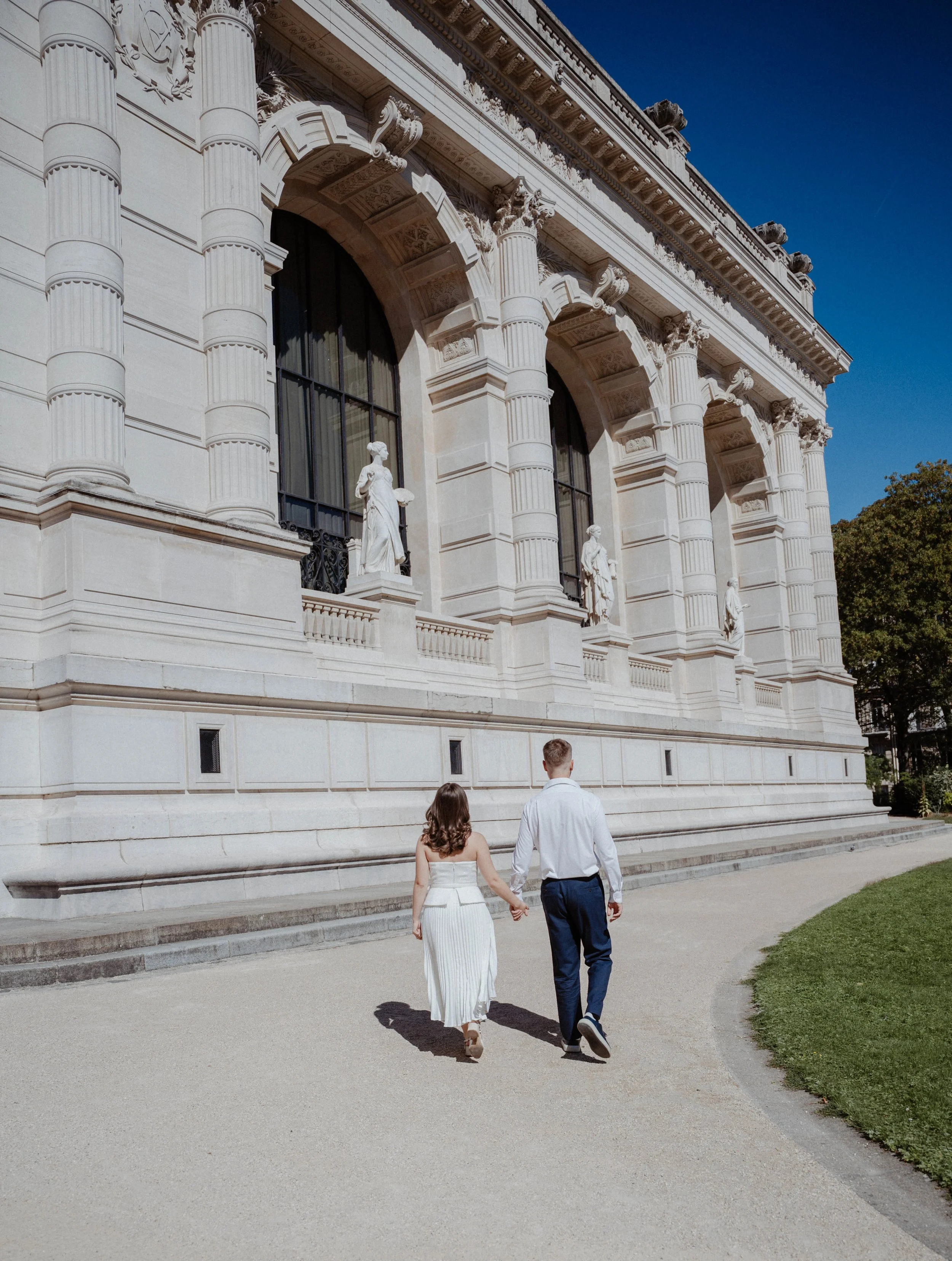 Un couple marche main dans la main devant un bâtiment ancien en pierre avec des colonnes et des statues décoratives, sous un ciel bleu.