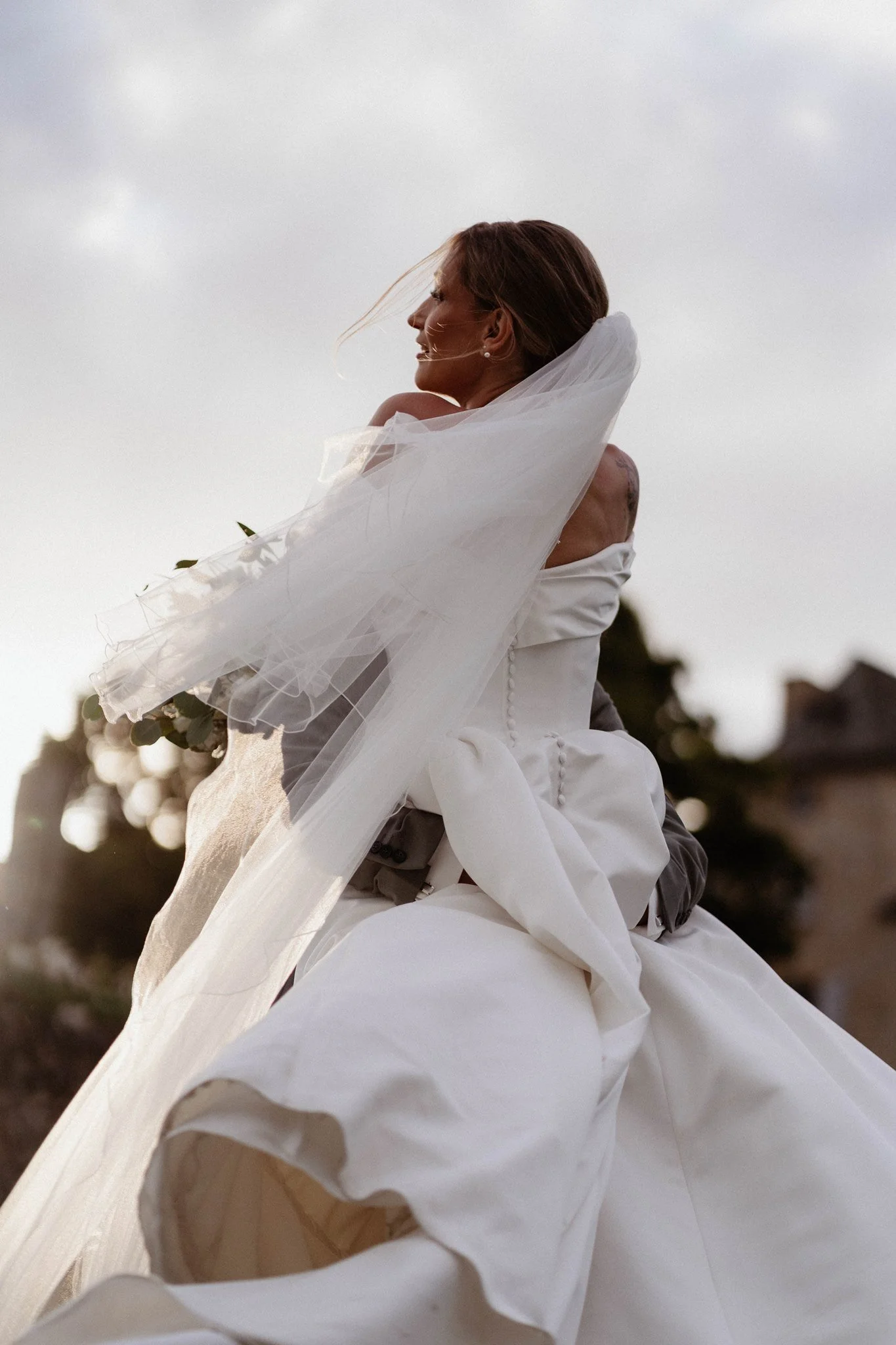 Une femme en robe blanche avec un voile, souriante, probablement lors d'un mariage, dans un décor en plein air avec un ciel nuageux.