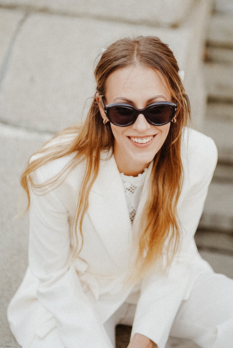 Femme souriante portant des lunettes de soleil noires, vêtue d'un costume blanc, avec des cheveux longs et roux, en extérieur