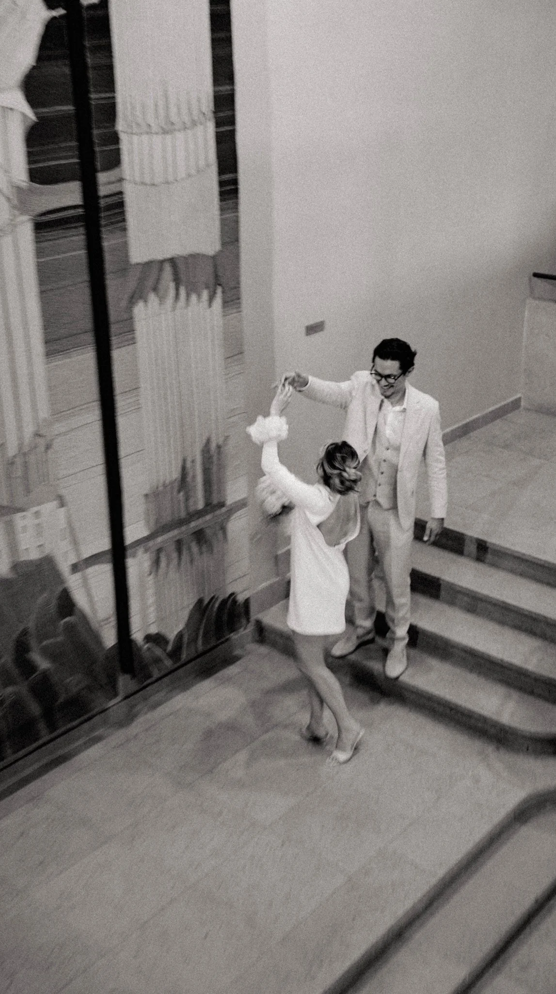 Deux personnes dansent dans une pièce avec un étage en bois. La femme porte une robe blanche et l'homme un costume clair. Il y a un mur blanc et un escalier en arrière-plan.