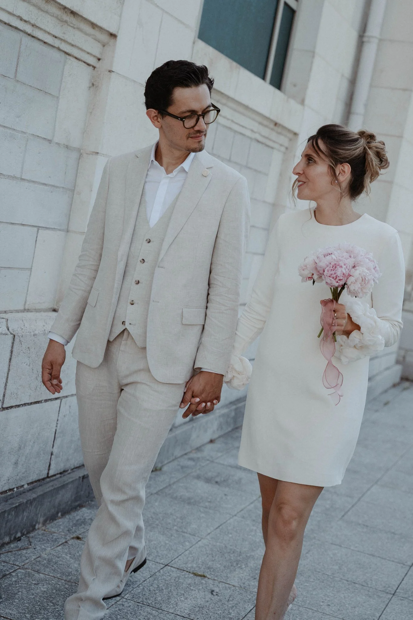 Un couple marié se tient la main en marchant dans la rue devant un bâtiment en pierre. La femme porte une robe blanche et tient un bouquet de roses, l'homme porte un costume beige clair.