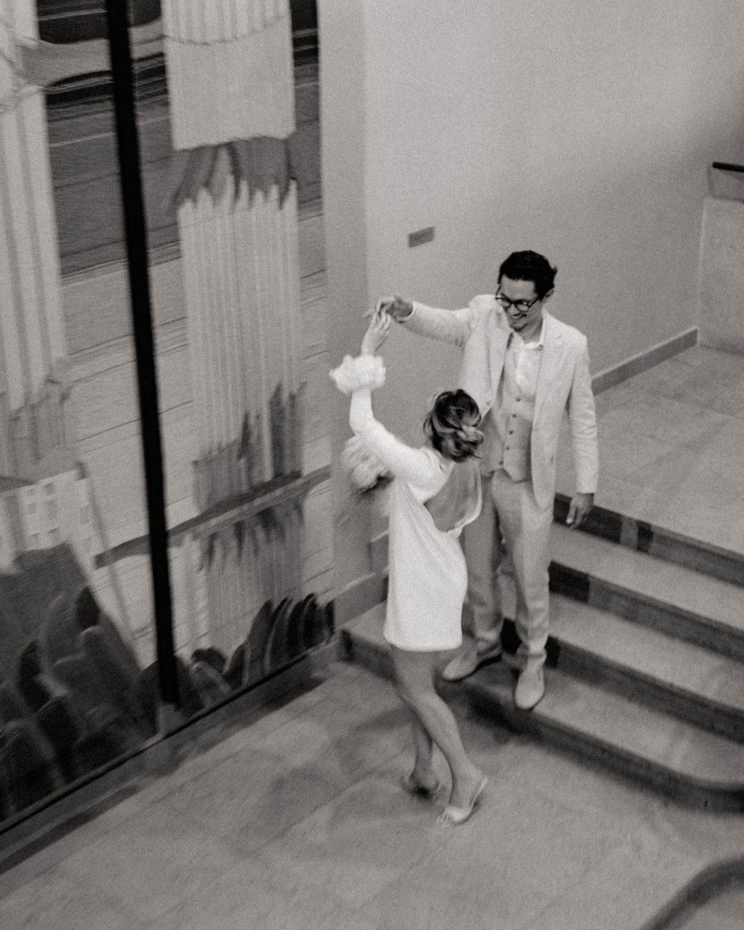 Un couple danse en haut d'un escalier dans une salle, l'homme porte un costume clair et la femme une robe blanche, photo en noir et blanc.