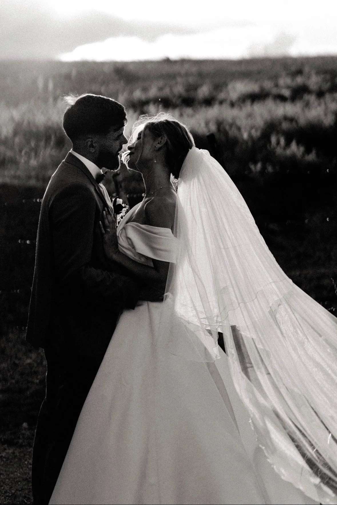 Un couple marié s'embrassant en noir et blanc, vêtus d'une robe de mariée et d'un costume, en plein air lors d'un coucher de soleil
