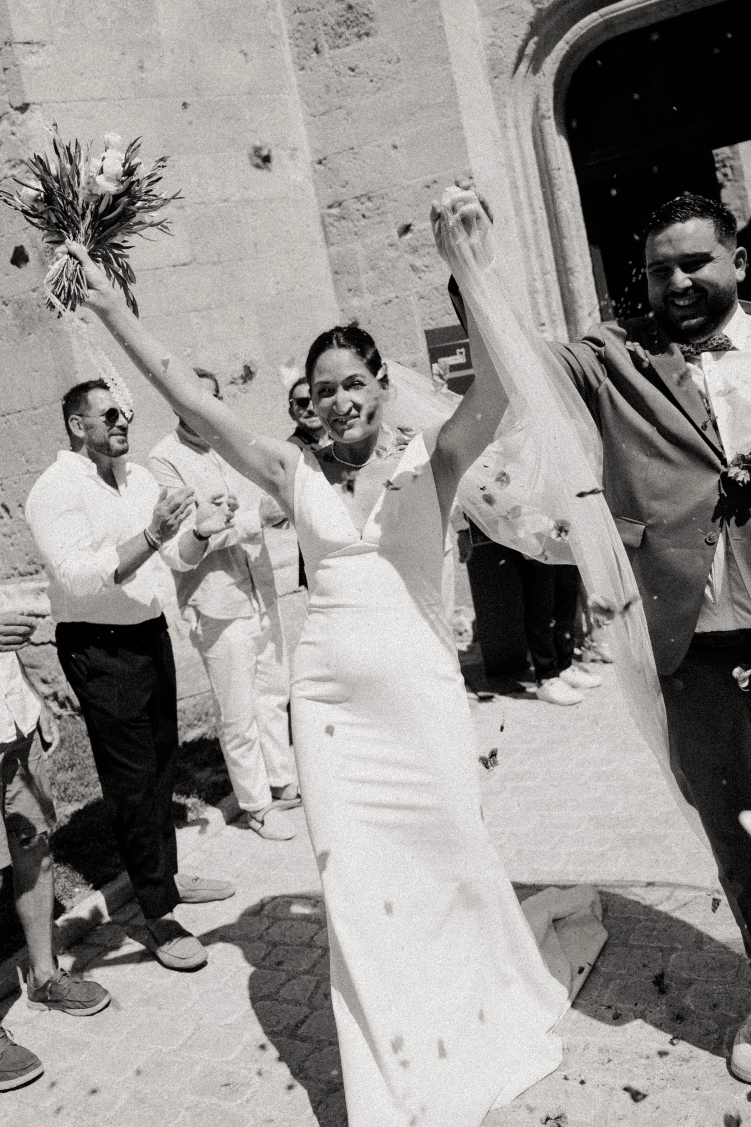 Une femme en robe de mariage et un homme en costume sourient lors d'une célébration en extérieur, accompagnés de plusieurs personnes.