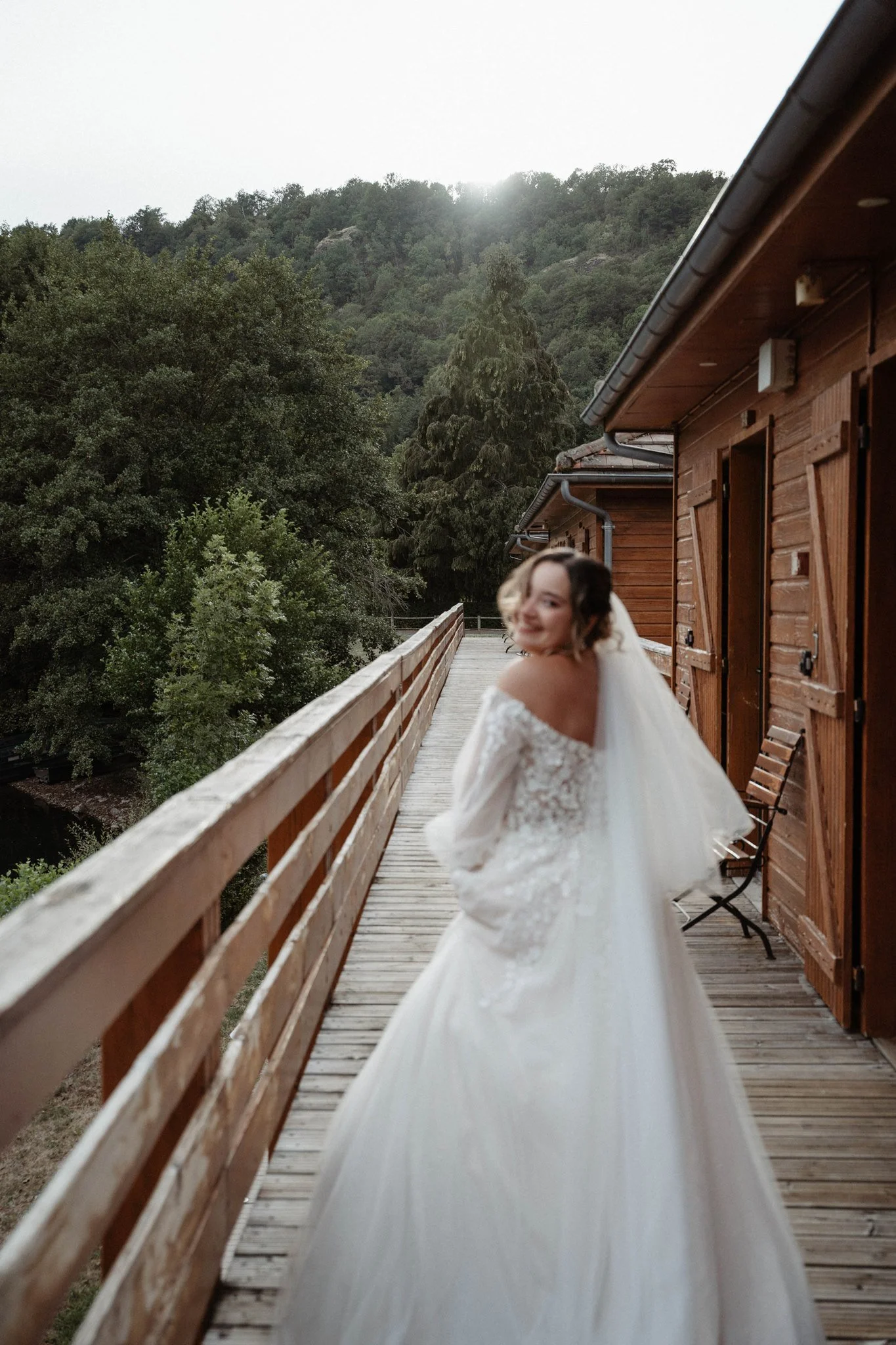 Une femme en robe de mariée sourit sur un balcon en bois en montagne avec des arbres verts et le soleil couchant en arrière-plan.