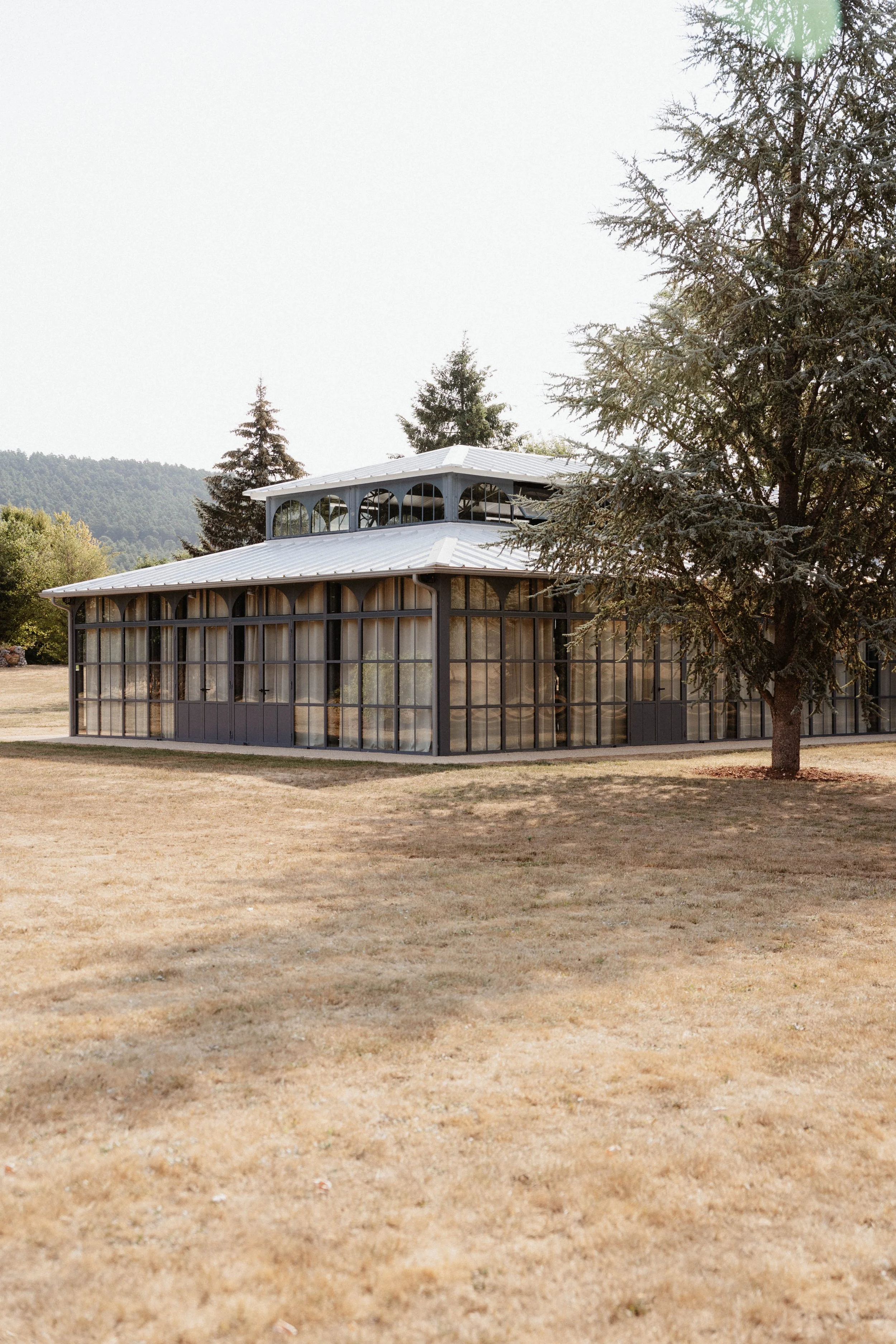 Une maison en verre avec un toit en métal, entourée d'arbres, dans un paysage rural avec une pelouse sèche