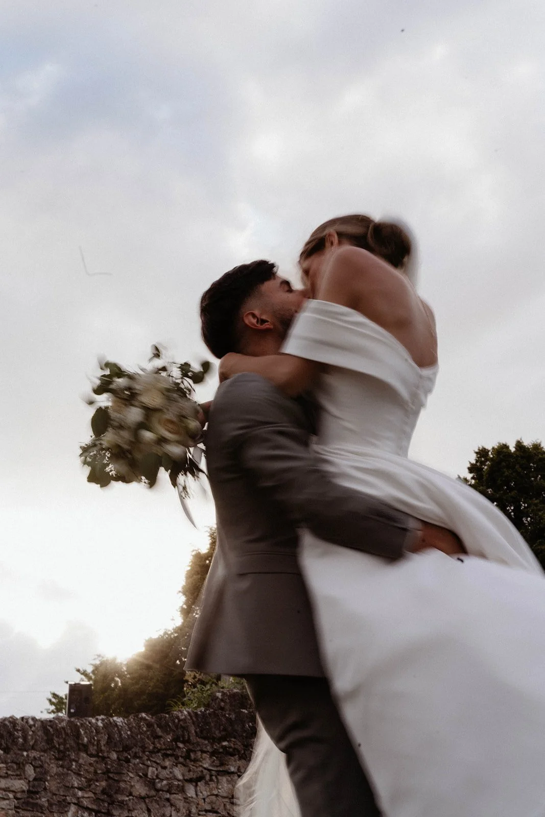 Un couple marié partage un moment romantique, l'homme portant la mariée dans ses bras, avec le ciel en arrière-plan.