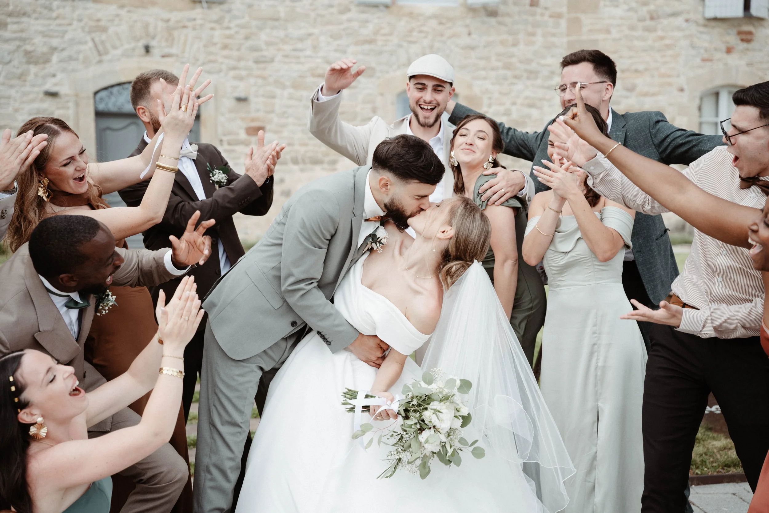 Un groupe de personnes en costume fête un mariage, avec un couple de mariés au centre qui s'embrassent, entourés de leurs amis qui les applaudissent et les encouragent.