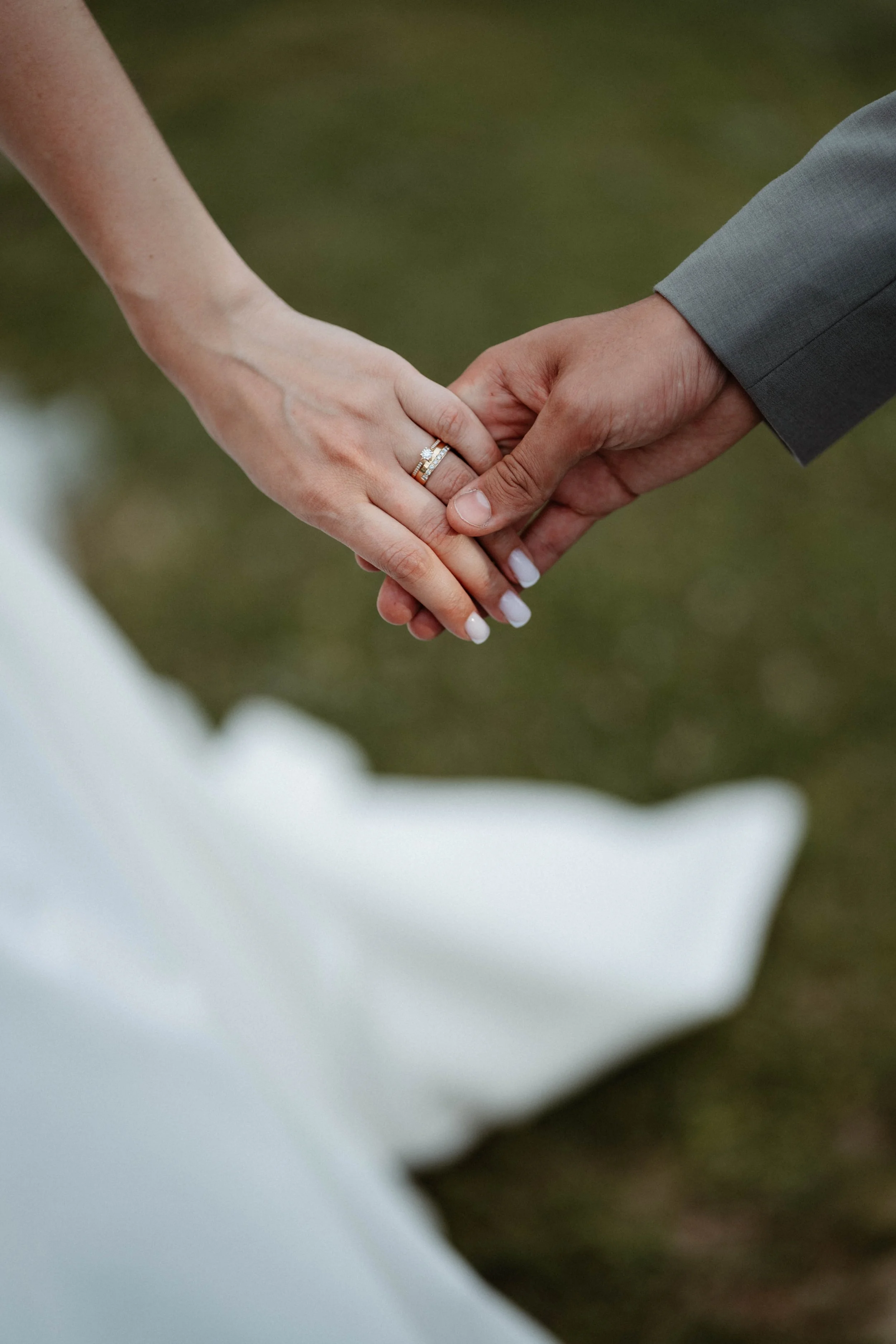 Le premier plan montre deux mains qui se tiennent, l'une portant une bague de mariage avec une pierre précieuse. La personne à gauche porte un vêtement blanc, probablement une robe de mariée, et la personne à droite porte une veste grise. L'arrière-p