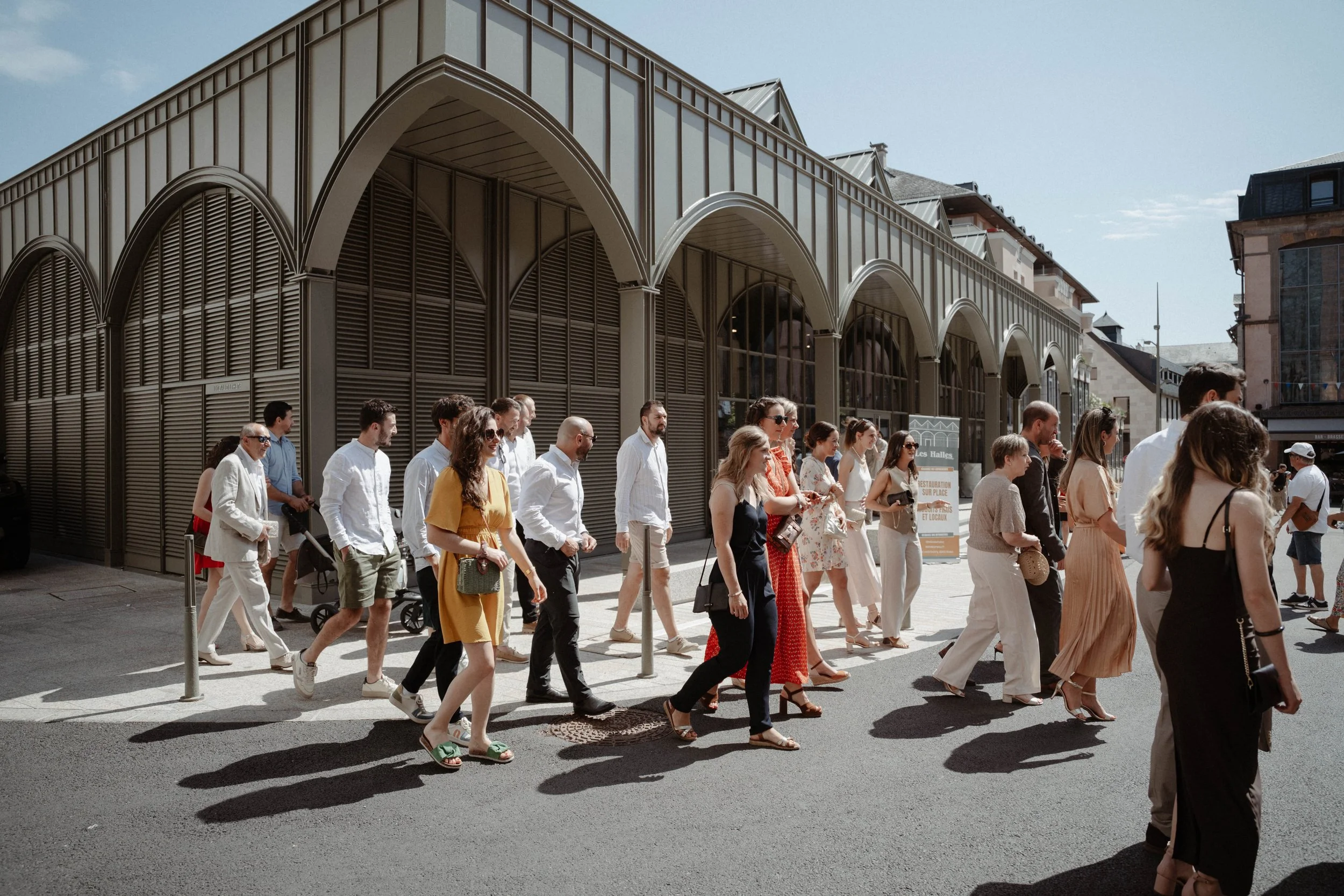 Groupe de personnes traversant la rue en plein jour devant un bâtiment moderne avec arches en métal et en verre.