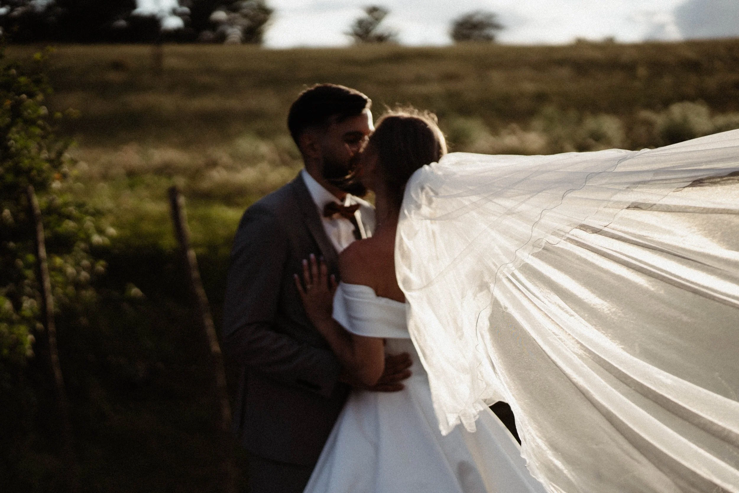 Un couple en tenue de mariage partage un baiser dans un champ rural avec un ciel partiellement nuageux.