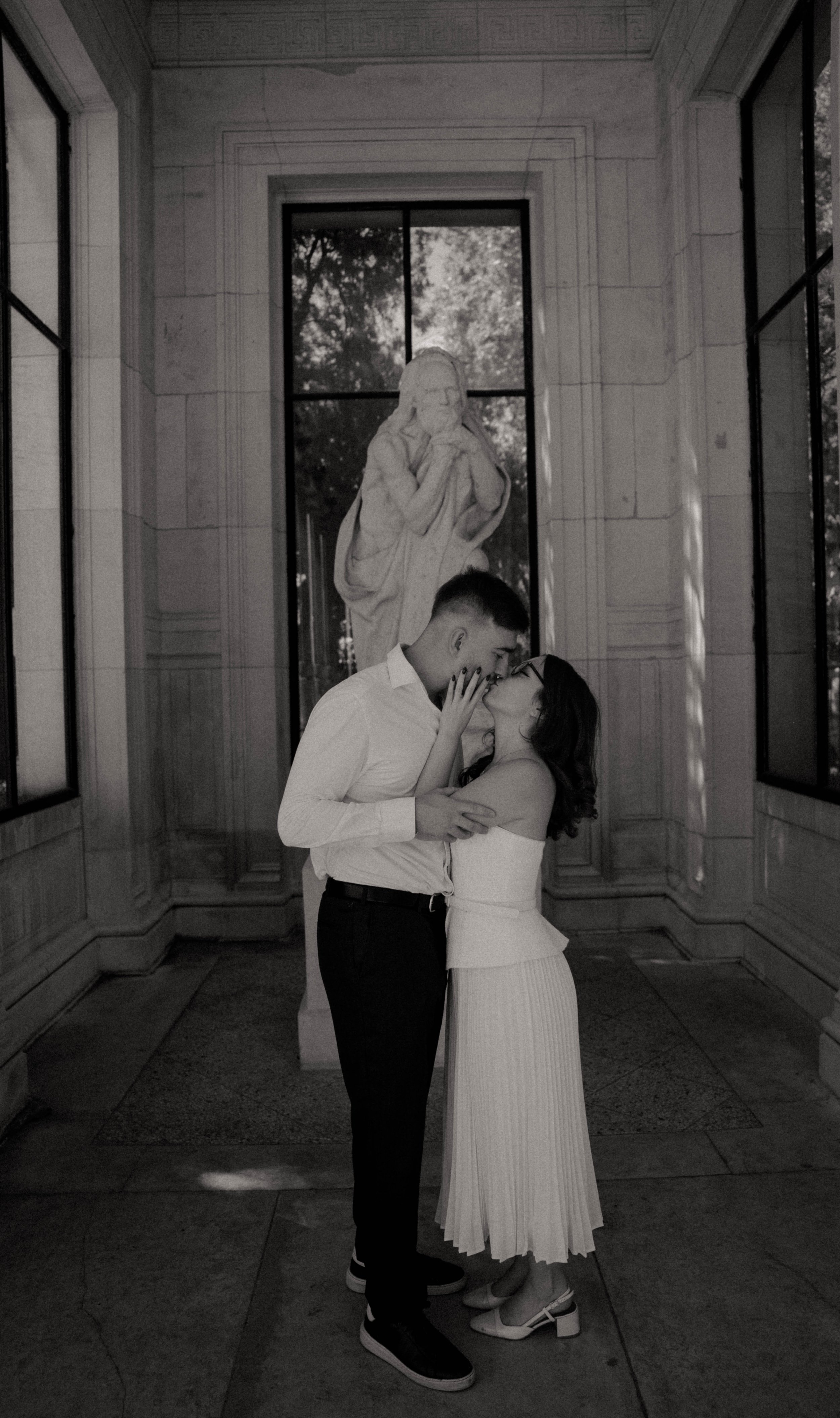 Un couple s'embrassant dans un couloir élégant avec des fenêtres et une statue en arrière-plan.