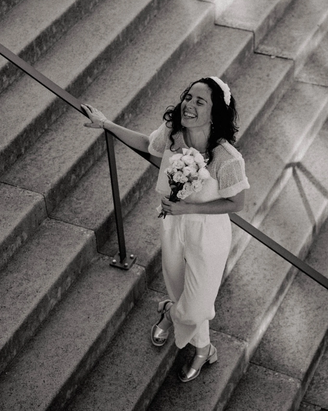 Une femme souriante en habits clairs tient un bouquet de fleurs et monte les escaliers en pierre, vue de dessus.