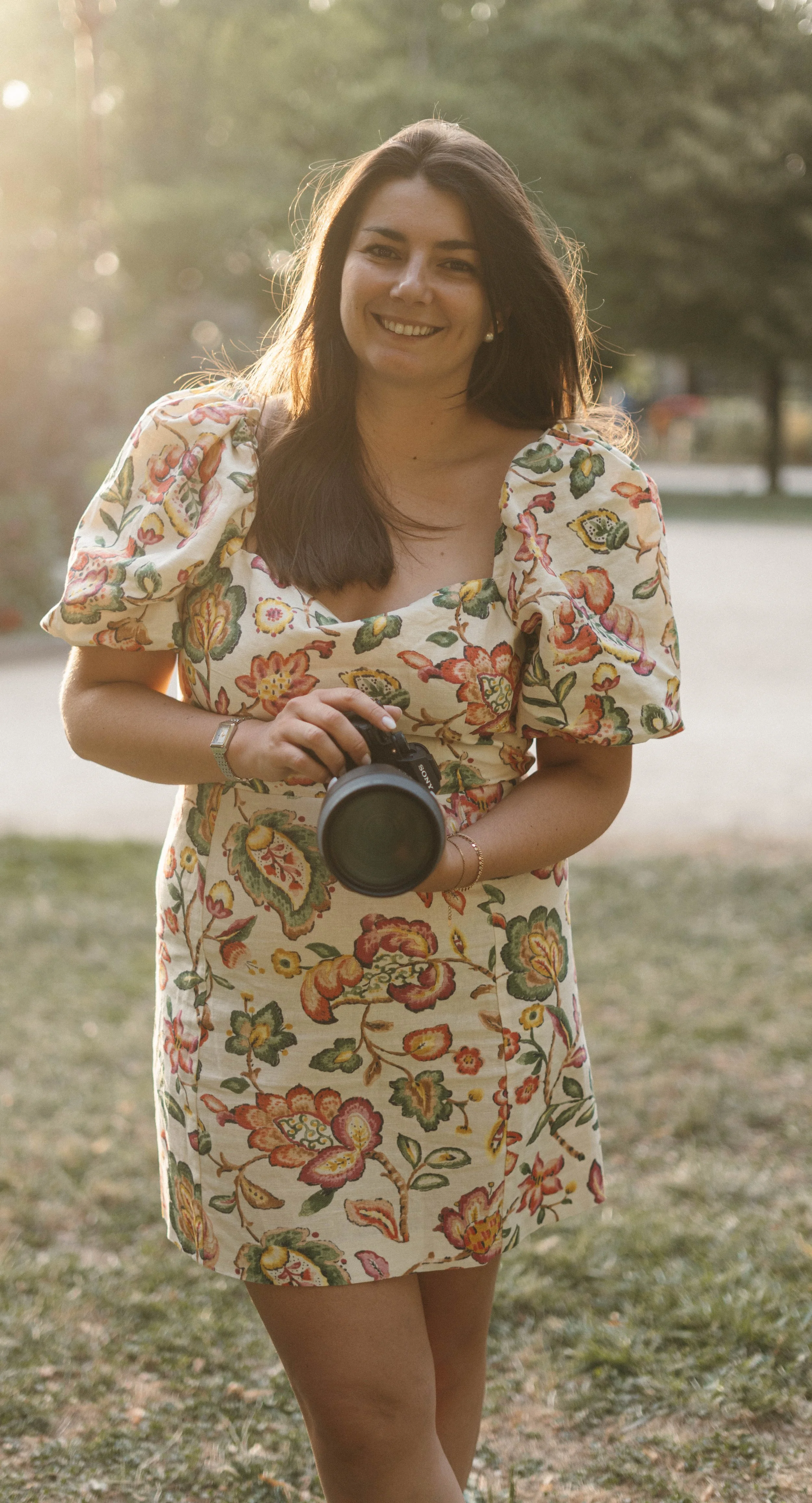 Femme souriante portant une robe à motifs floraux, tenant un appareil photo en plein air au coucher du soleil.