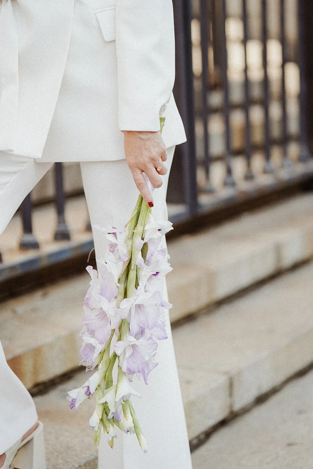 Une personne en costume blanc tenant un bouquet de fleurs blanches, près d'une grille en métal, probablement dans un cadre urbain ou un espace public.