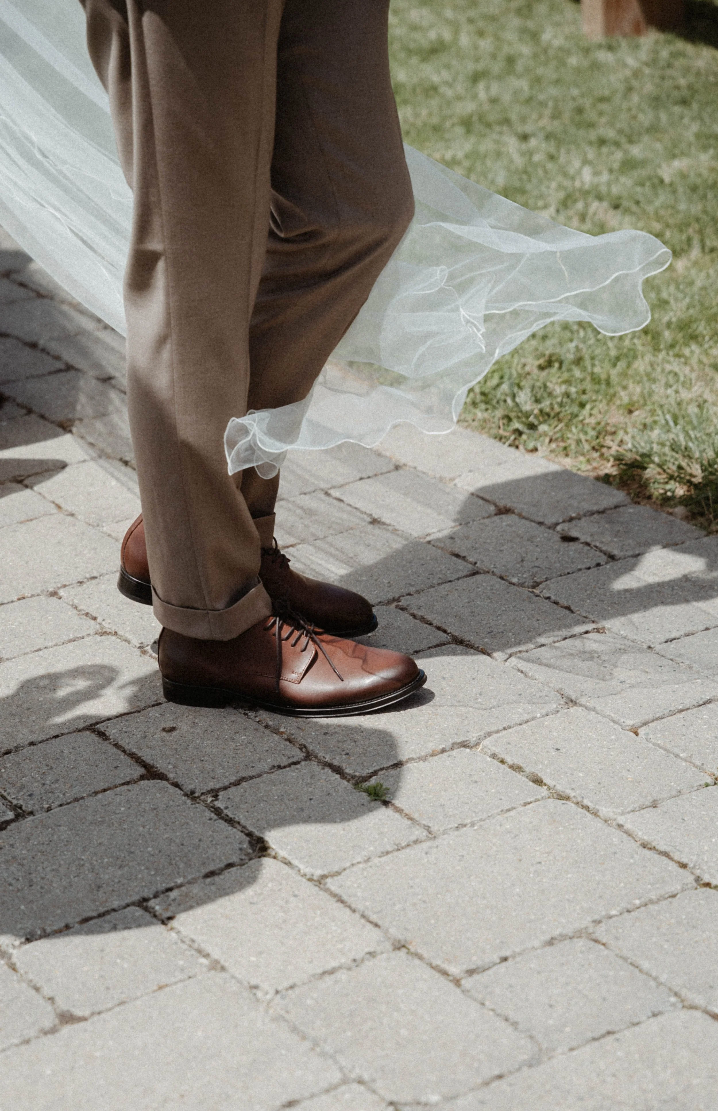 Les jambes d'une personne portant un pantalon marron, des chaussures en cuir marron et une jupe en tissu transparent, marchant sur un trottoir pavé avec de l'herbe à côté.