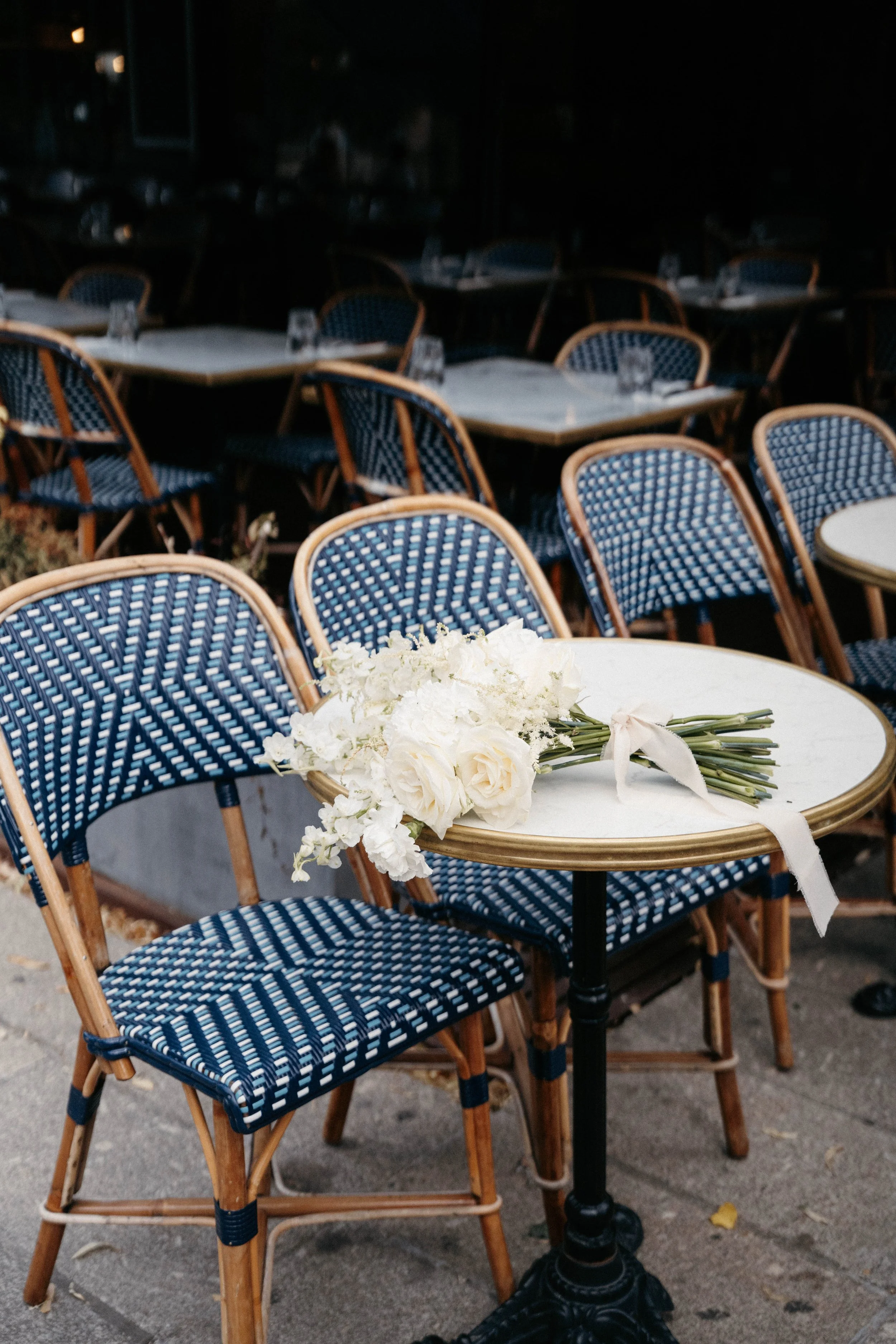 Une femme portant un pull blanc et un pantalon à rayures, tenant un bouquet de fleurs blanches dans une boutique de fleurs avec des étagères contenant des verres, des vases en porcelaine, et des plantes vertes