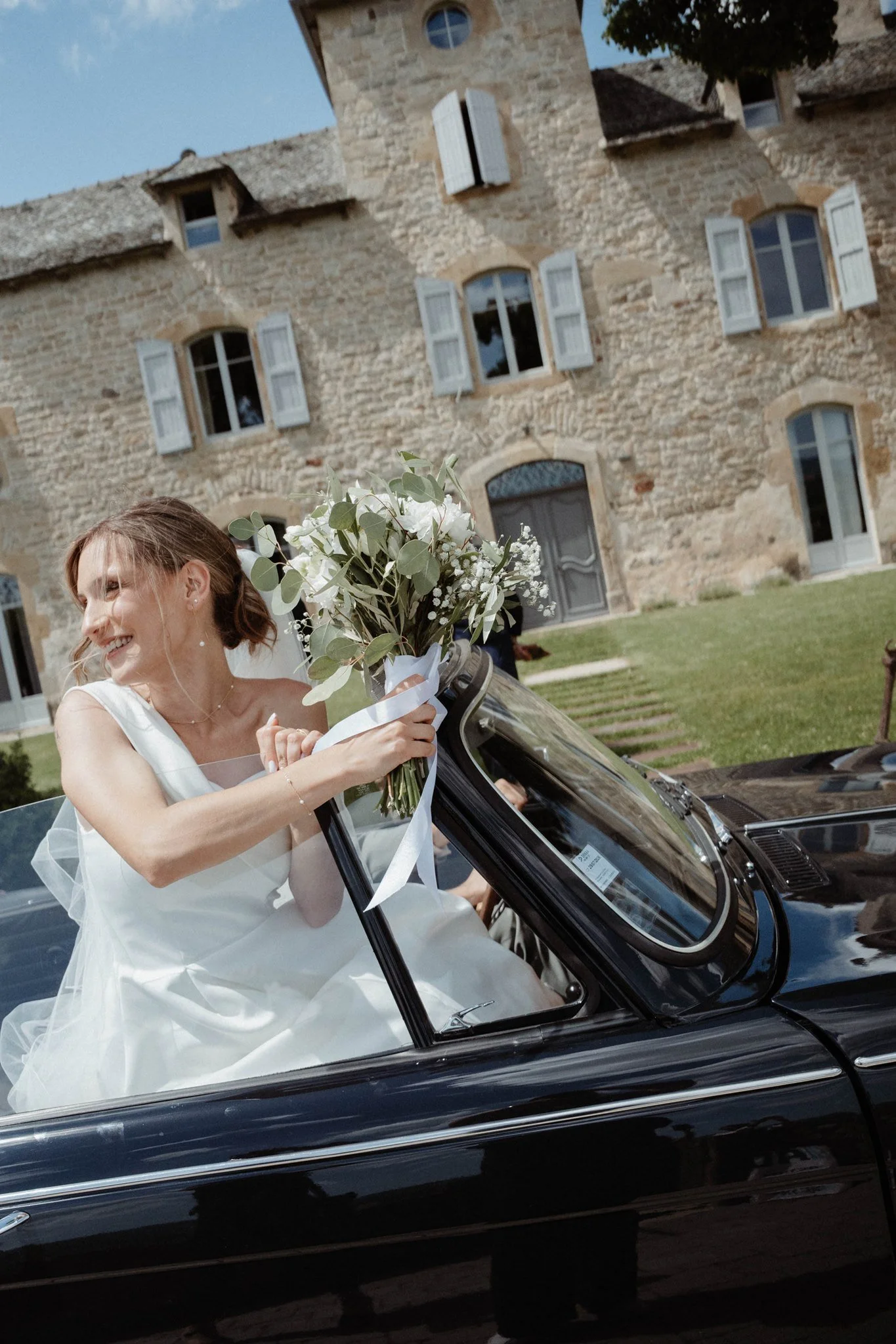 Une femme en robe de mariage sourit en tenant un bouquet de fleurs blanches, assise dans une voiture noire de luxe, devant un bâtiment en pierre.