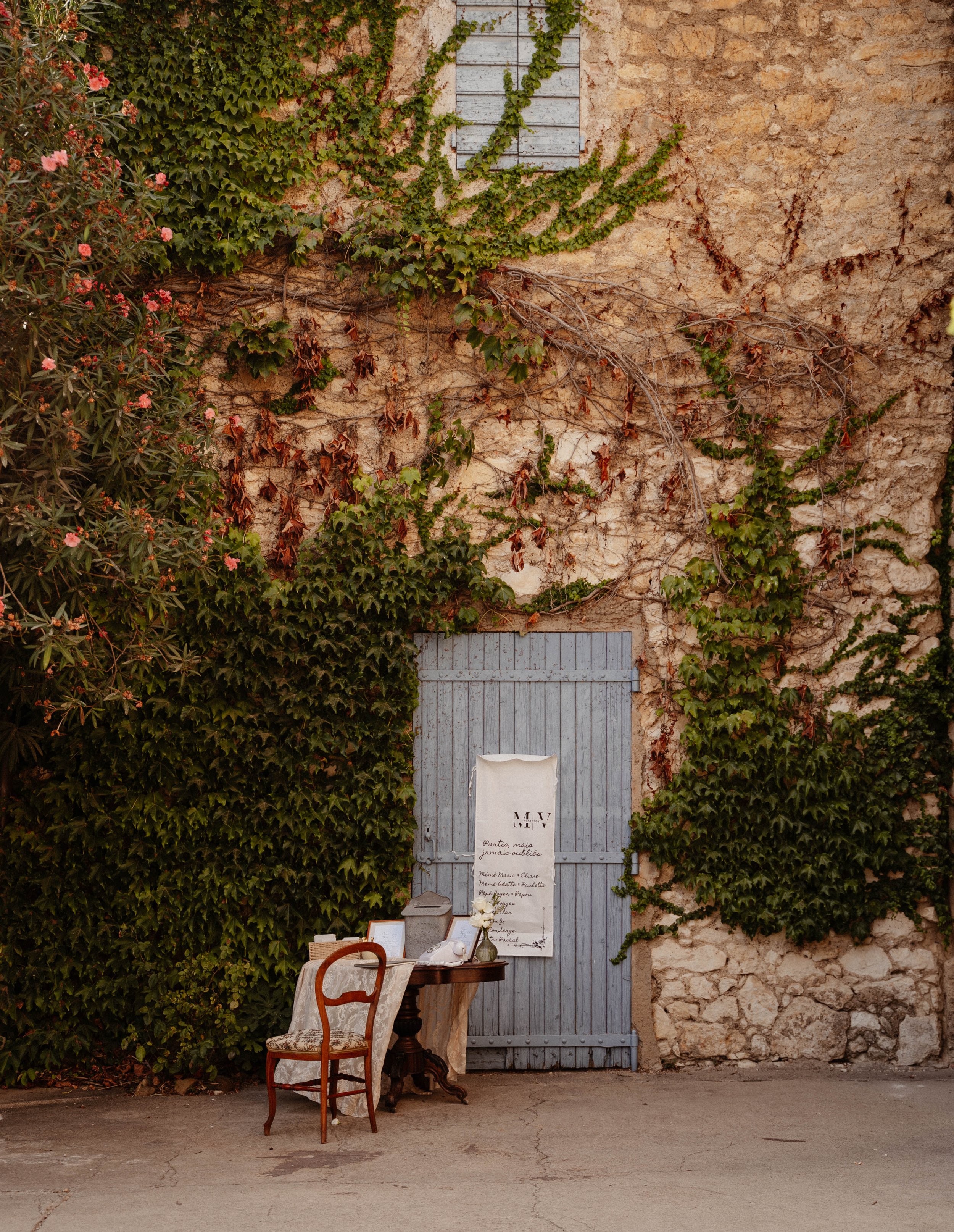 Une porte en bois grise située sur un mur en pierre, entourée de vignes et de plantes à feuilles vertes et marron. Devant la porte, une table avec deux chaises, une nappe blanche et des papiers ou menus.