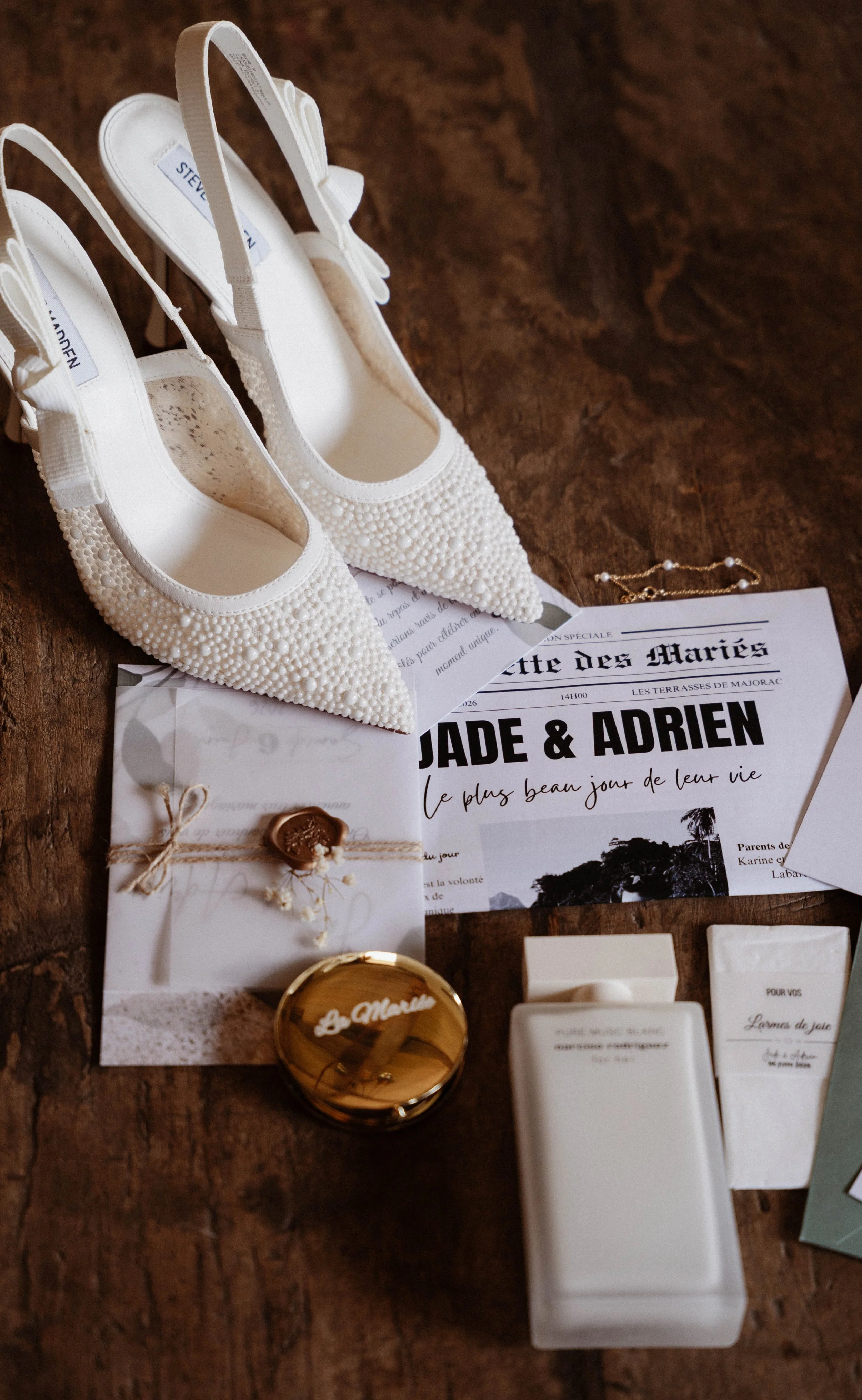 Une paire de chaussures de mariage blanches ornées de perles et une petite enveloppe scellée avec de la cire et une corde en jute, posées sur une surface en bois, entourées de diverses cartes et objets de décoration