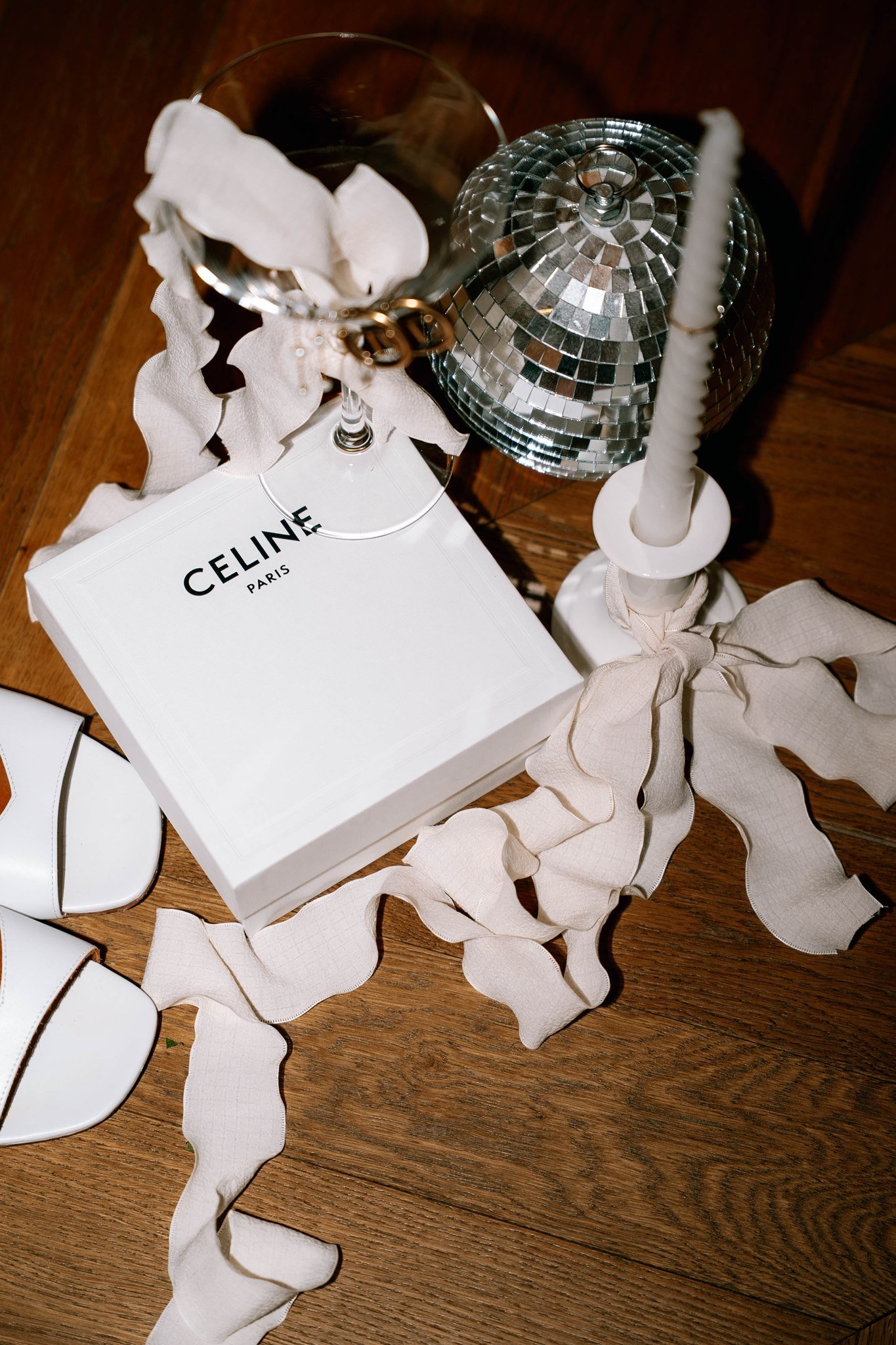 Une boîte blanche avec le texte 'CELINE PARIS' est posée sur une table en bois. Autour, il y a un verre à vin, un chandelier blanc et une décoration de miroir en mosaïque. Des rubans en tissu beige décorent la scène.