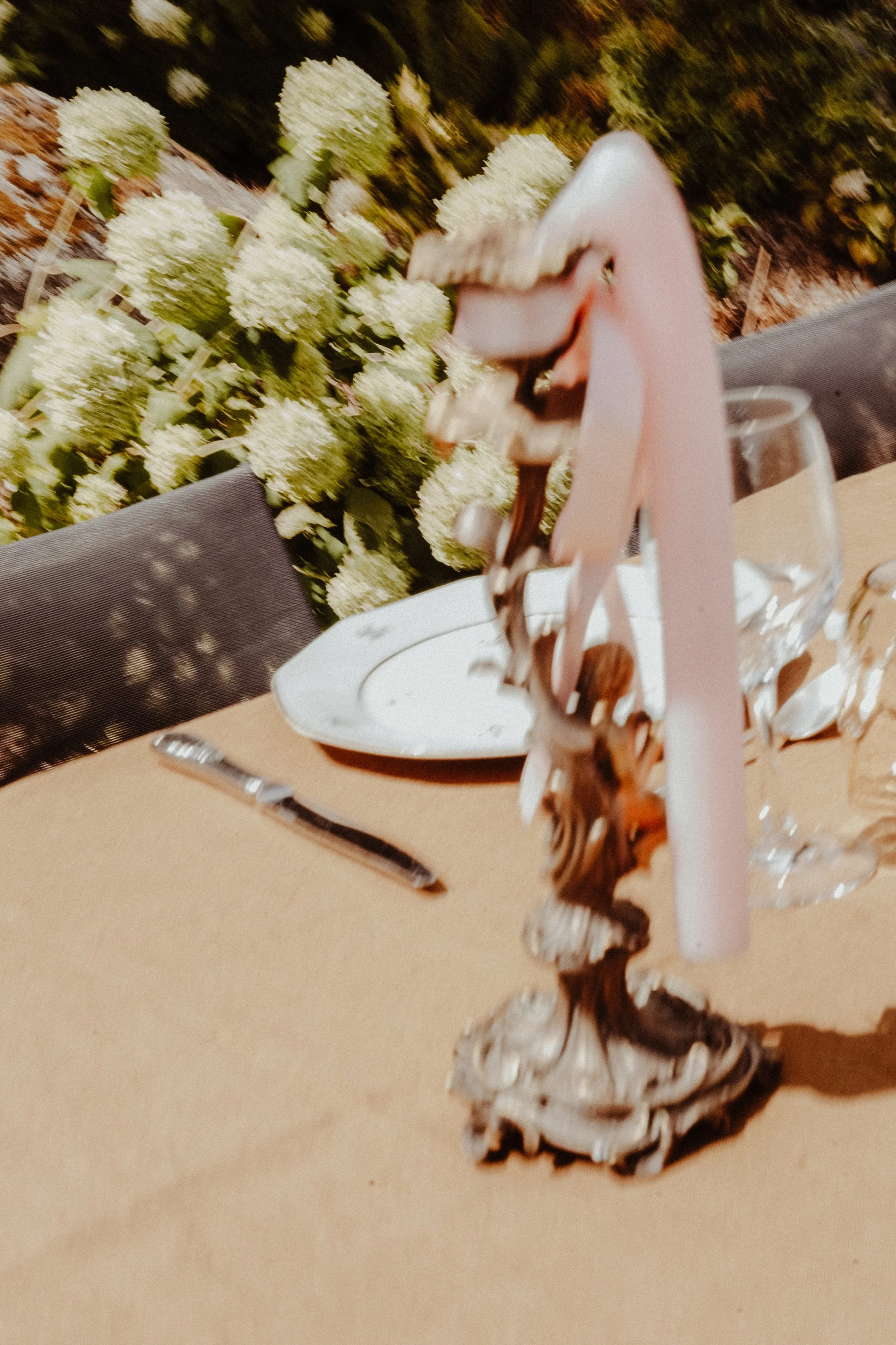 Une décoration de table avec un centre de table en forme de spirale, une assiette blanche, un verre à pied et une fourchette sur une nappe beige. En arrière-plan, un bouquet de fleurs blanches et une toile de fond verte