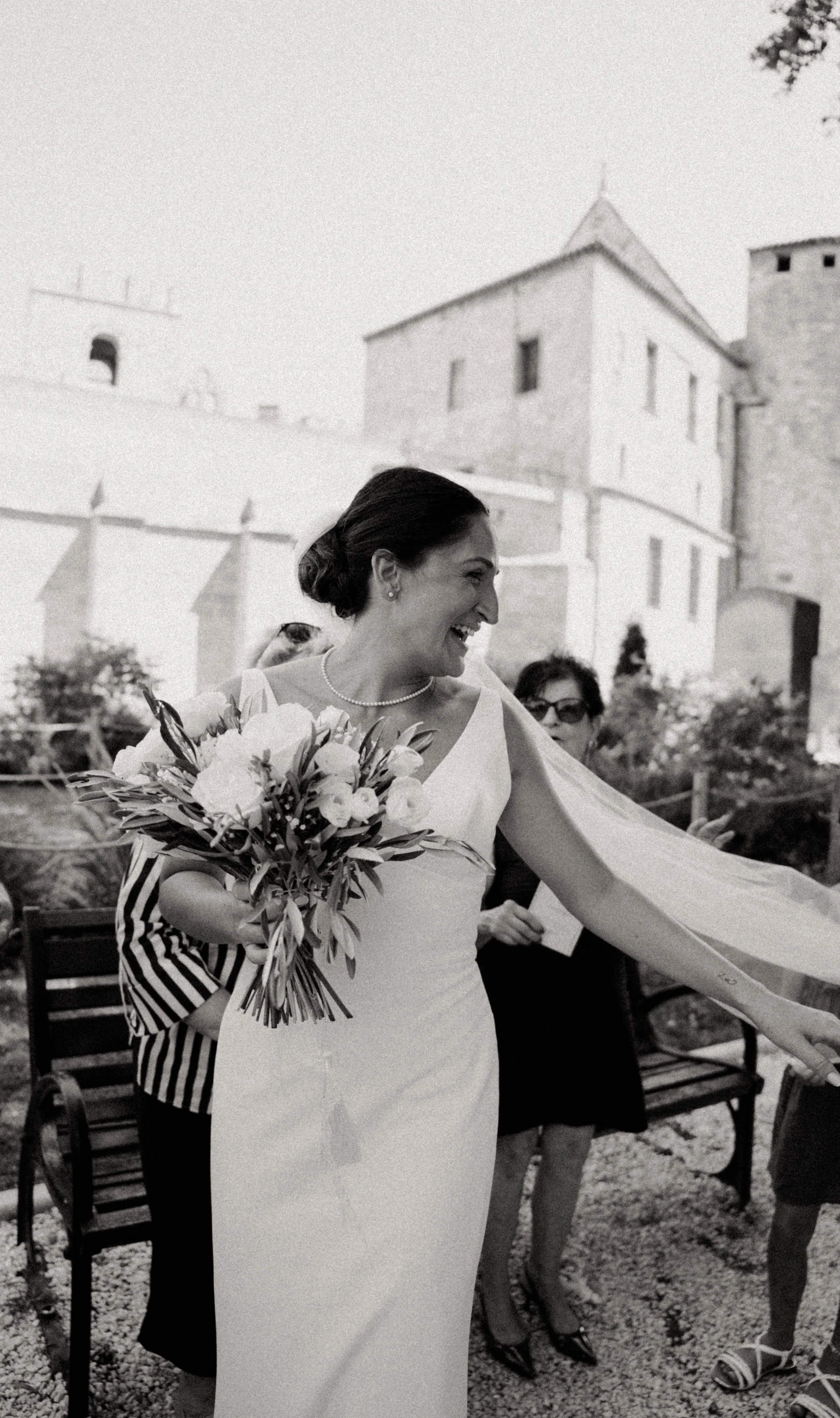 Une femme souriante portant une robe blanche et tenant un bouquet de fleurs, lors d'un événement en plein air, avec des personnes autour et un château en arrière-plan.