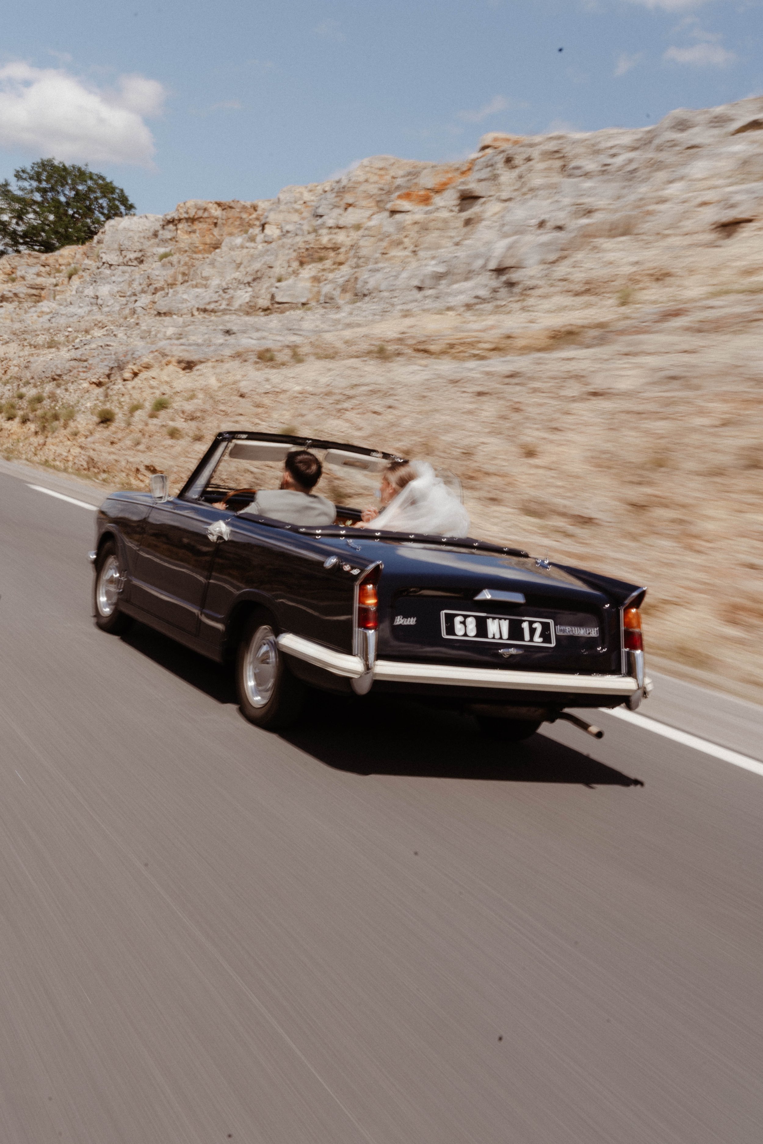 Une voiture classique noire, conduite par un homme, transportant un couple dont la femme porte une robe de mariée, en déplacement sur une route à travers un paysage rocheux sous un ciel partiellement nuageux.