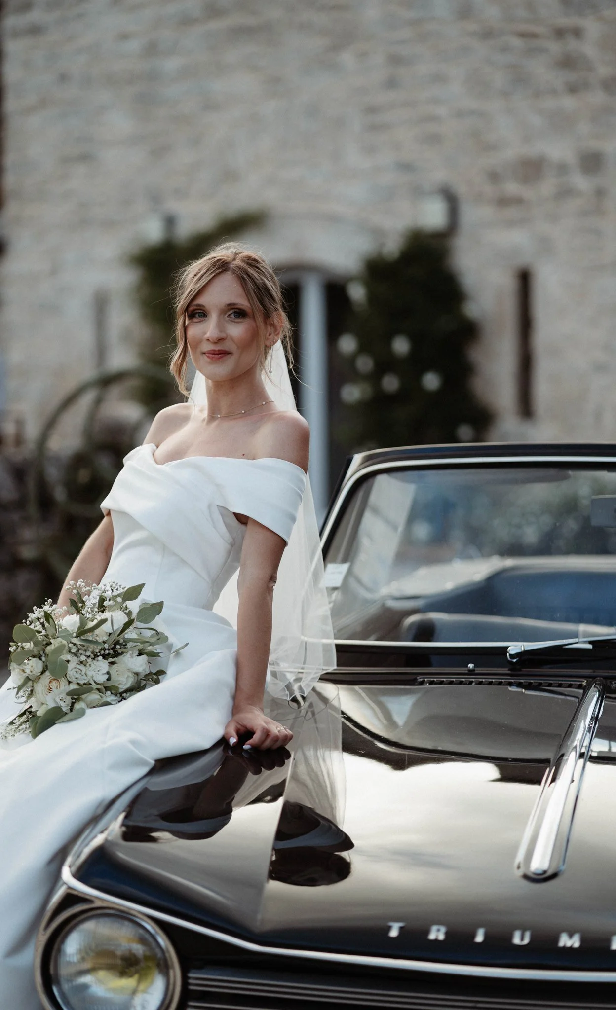 Une femme en robe de mariée blanche assise sur le capot d'une voiture de style vintage noire, tenant un bouquet de fleurs, dans une cour en pierre avec un mur en arrière-plan.