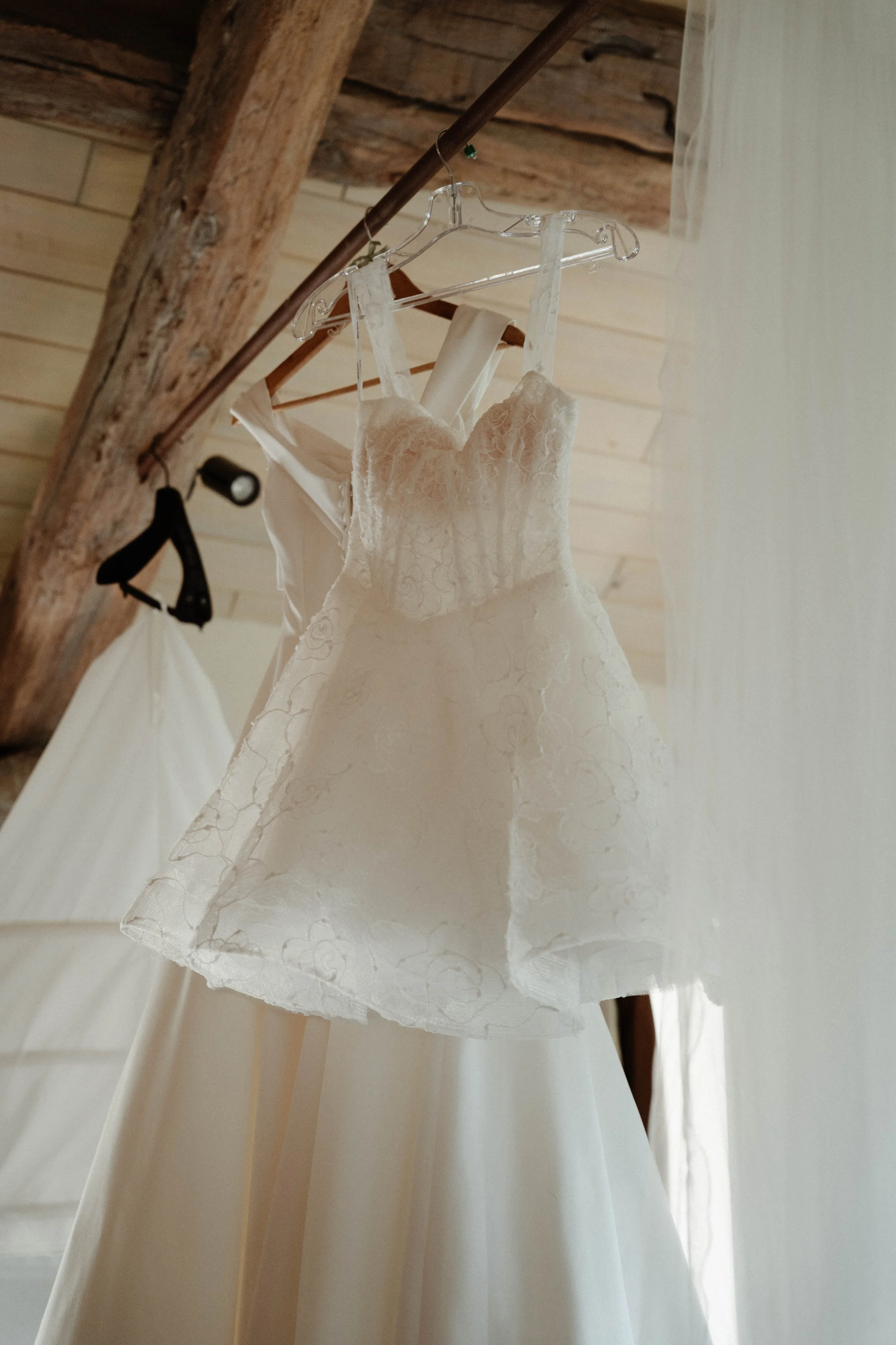 Robe de mariée blanche suspendue, en tissu avec des détails en dentelle, accrochée à un cintre en plastique, dans un espace avec plafond en bois.
